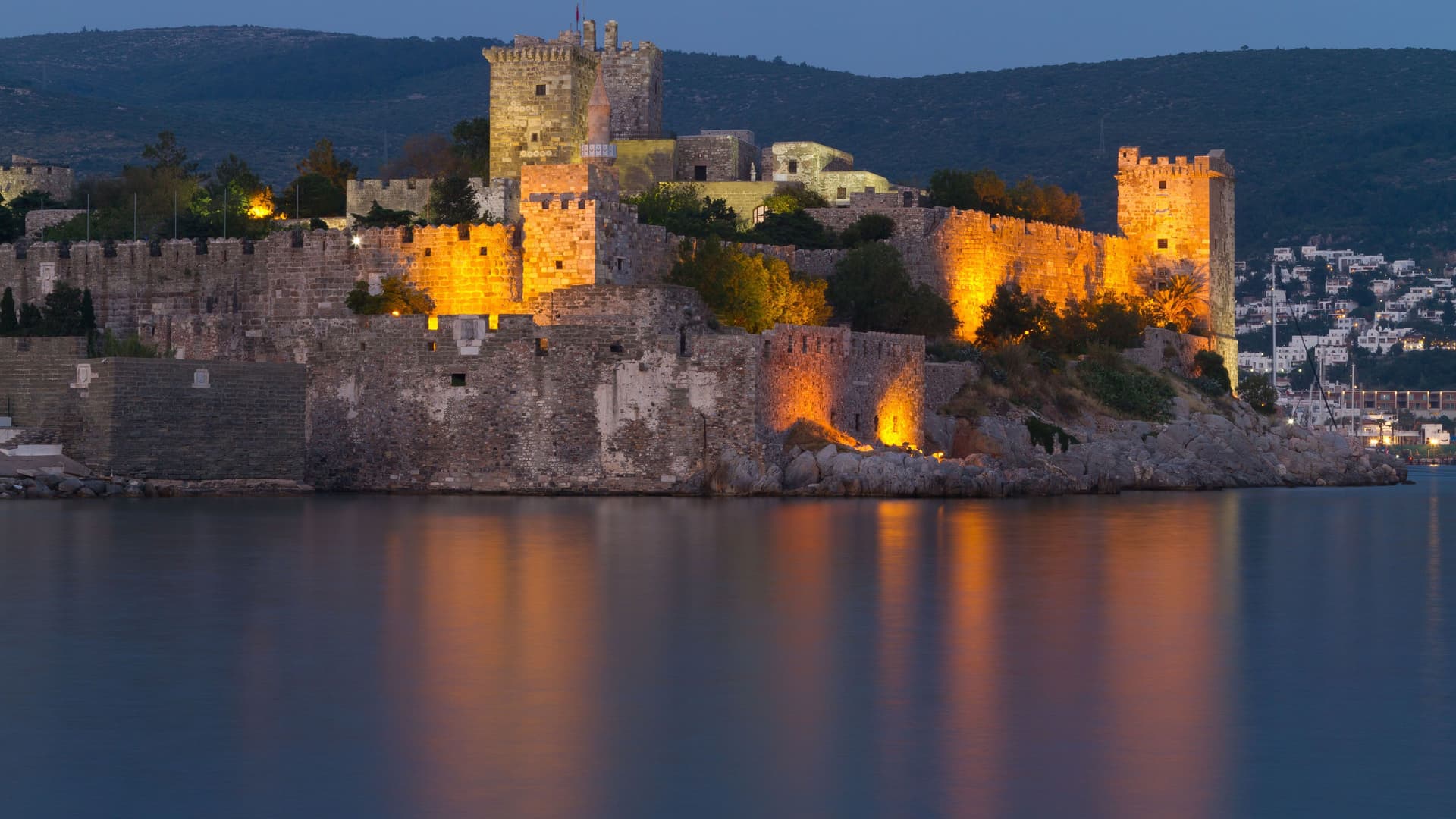 A dramatic dusk shot of the historic Bodrum Castle, also known as the Castle of St. Peter, illuminated at night, with its reflection visible in the calm waters of the harbor in Turkey.