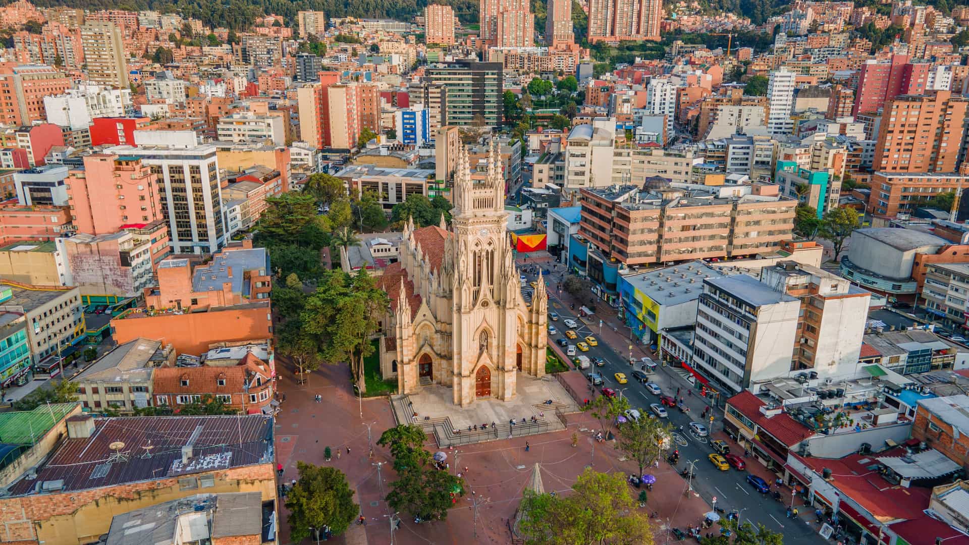 An aerial view of Bogota, Colombia, centered on the historic Church of Our Lady of Lourdes, a Gothic-style cathedral with twin spires. The church is surrounded by a large public square and a busy street filled with traffic. In the background, the city's densely packed buildings stretch up a green, forested hillside.