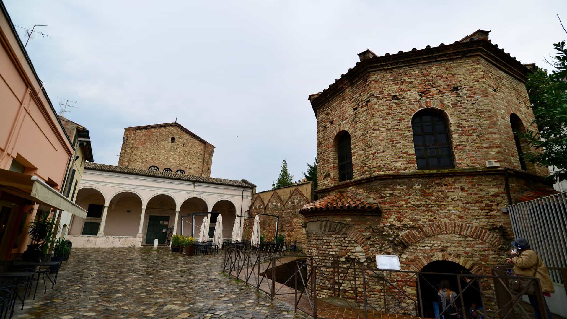 A view of an historic cobblestone courtyard in Ravenna, Italy, under an overcast sky. On the right is a prominent, octagonal, red-brick building with arched windows, which is the Baptistery of Neon. To the left is a building with a covered walkway of arches, and in the foreground are tables and chairs from a cafe.