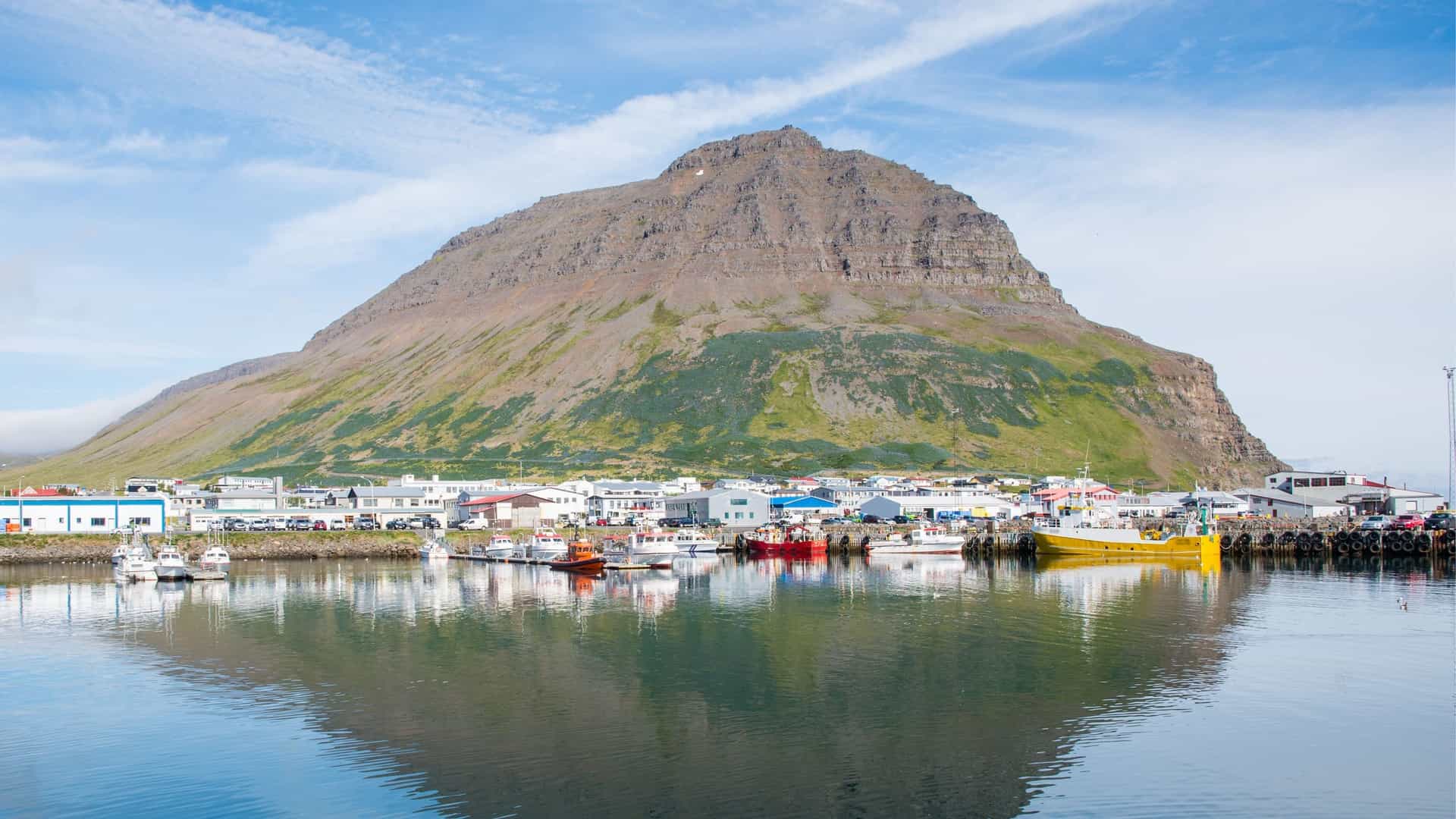 A panoramic view of the harbor in Bolungarvik, Iceland, showing fishing boats docked along the pier with a quaint town at the base of a massive, imposing mountain reflected in the still water.