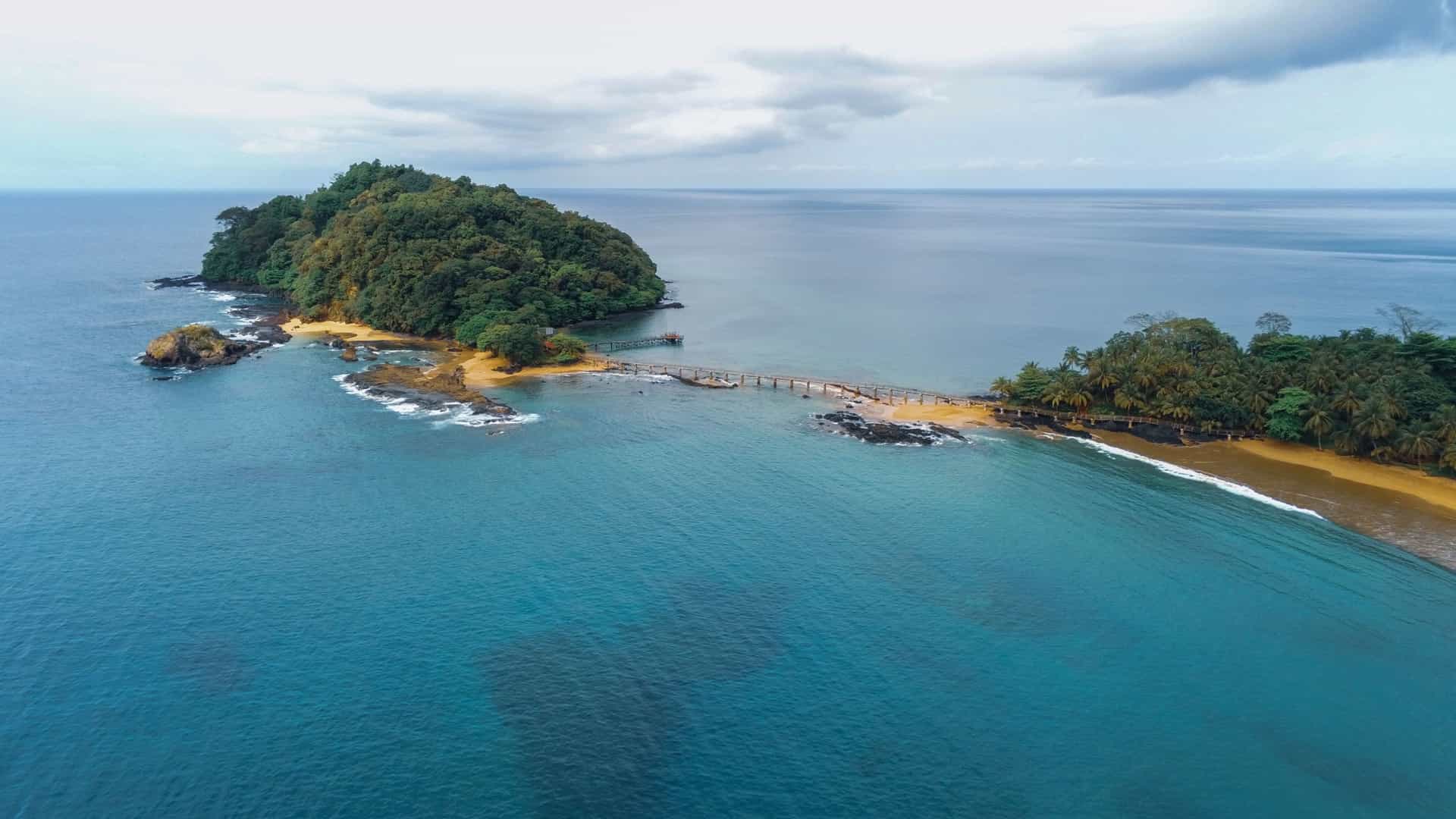 An aerial shot of Bom Bom Island, Sao Tome and Principe, showing the small, lushly forested island resort connected by a long wooden walkway to the mainland's sandy beach.