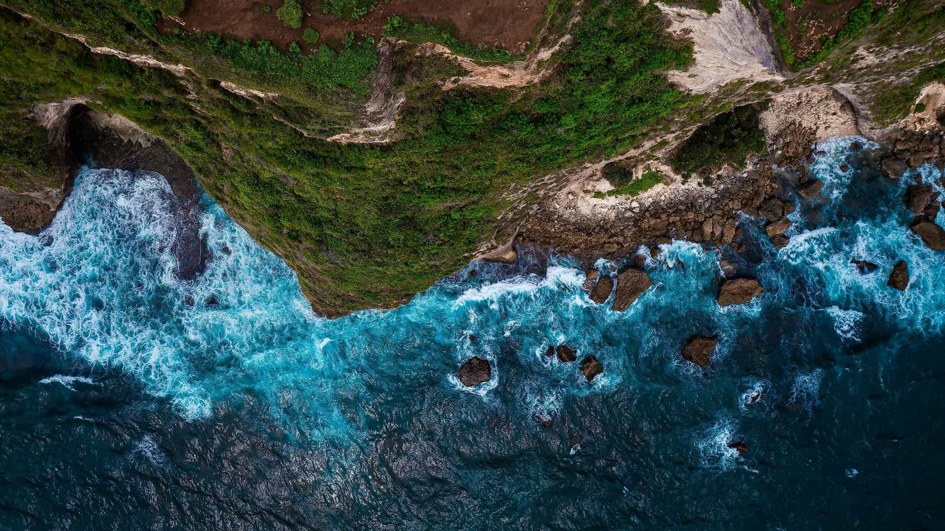 An aerial view of a rugged, green cliffside in Boma, Democratic Republic of Congo, showing powerful turquoise waves crashing against the rocky shoreline below.