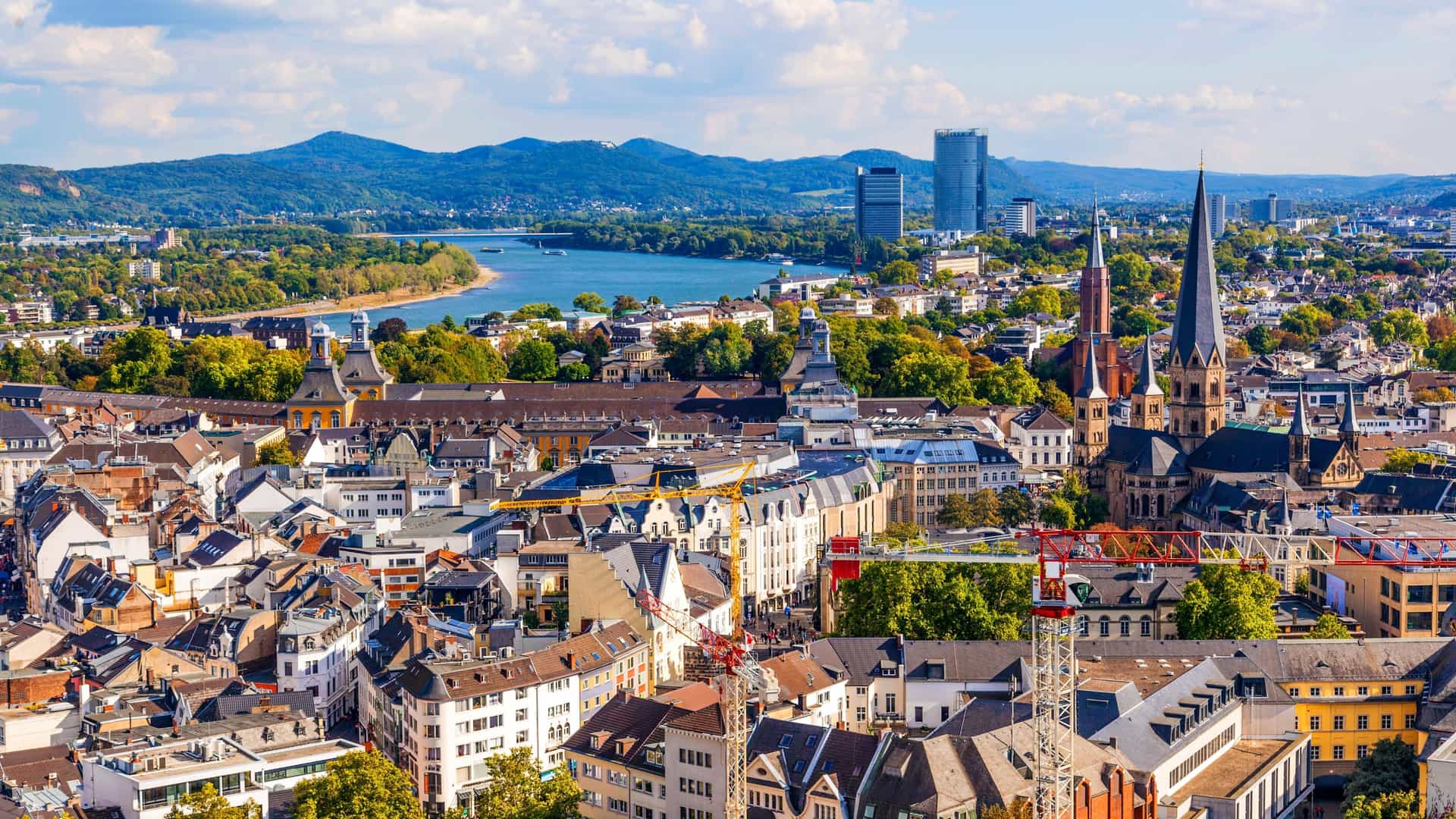 A panoramic, elevated view of Bonn, Germany, showing the urban area, the Rhine river, and the surrounding distant hills under a sunny, blue sky.