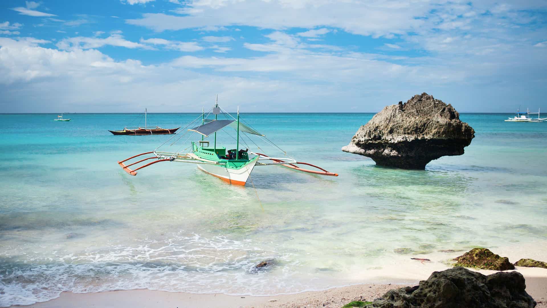 A vibrant outrigger boat floats in the crystal-clear, turquoise waters of Boracay Island in the Philippines, with a unique rock formation in the background.