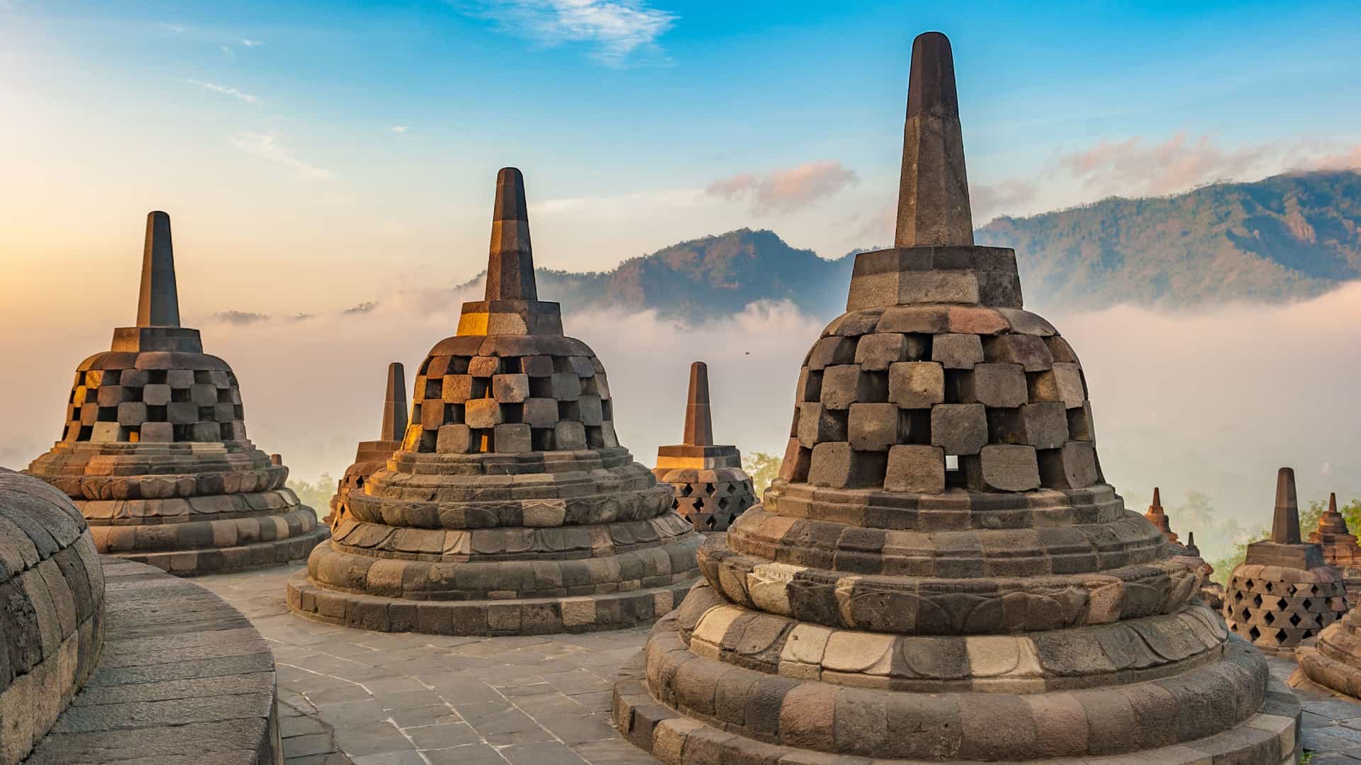 A view of the upper terraces of the Borobudur Buddhist temple in Indonesia at sunrise. In the foreground, several large, gray, bell-shaped stupas with open-worked lattices are visible on a stone platform. The background features a landscape of misty, forested mountains under a sky with soft orange, pink, and blue hues.