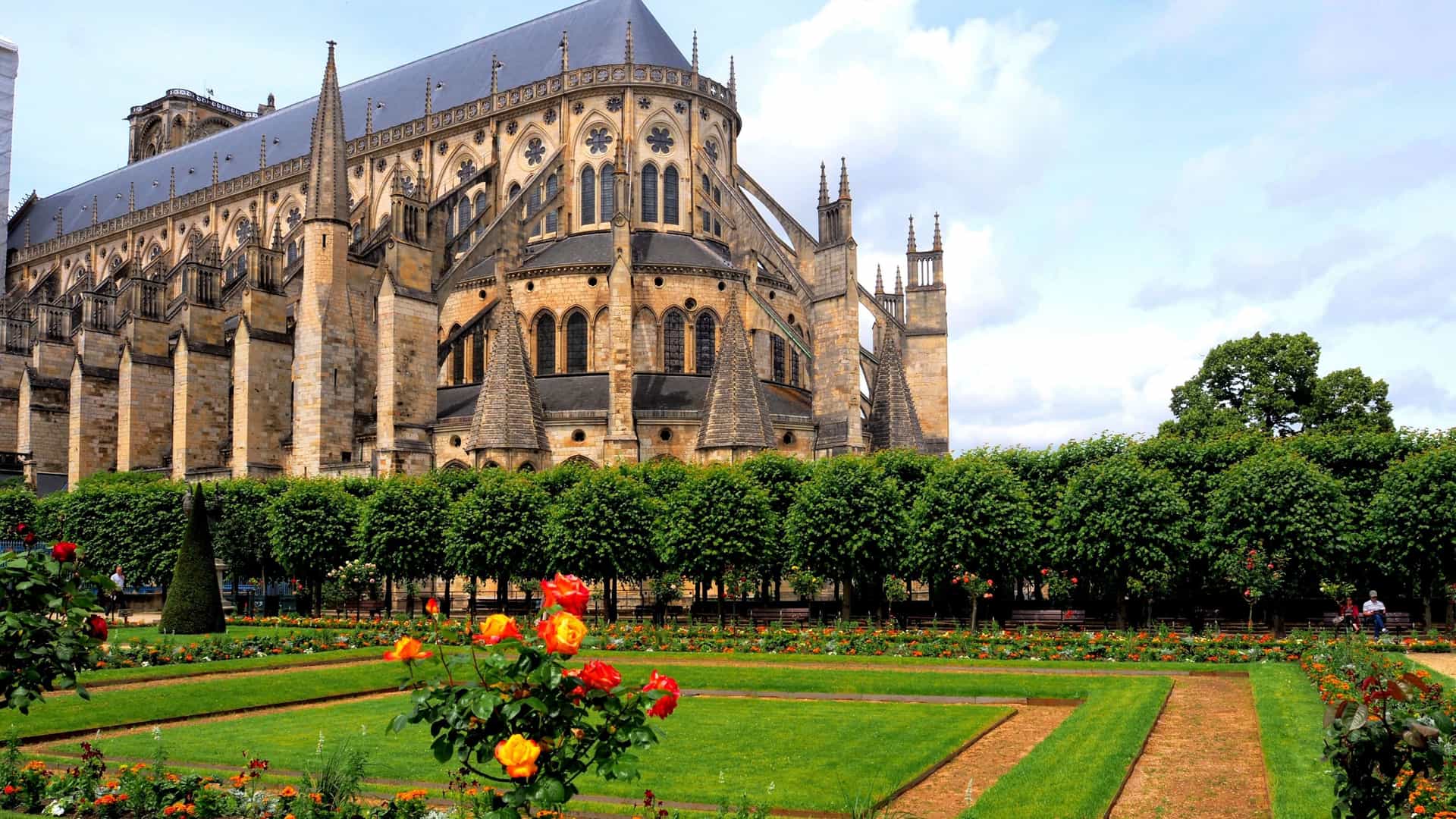 A view of the historic Saint-Étienne Cathedral in Bourges, France, showcasing the intricate Gothic architecture from a beautifully landscaped garden with manicured hedges and bright flowers.