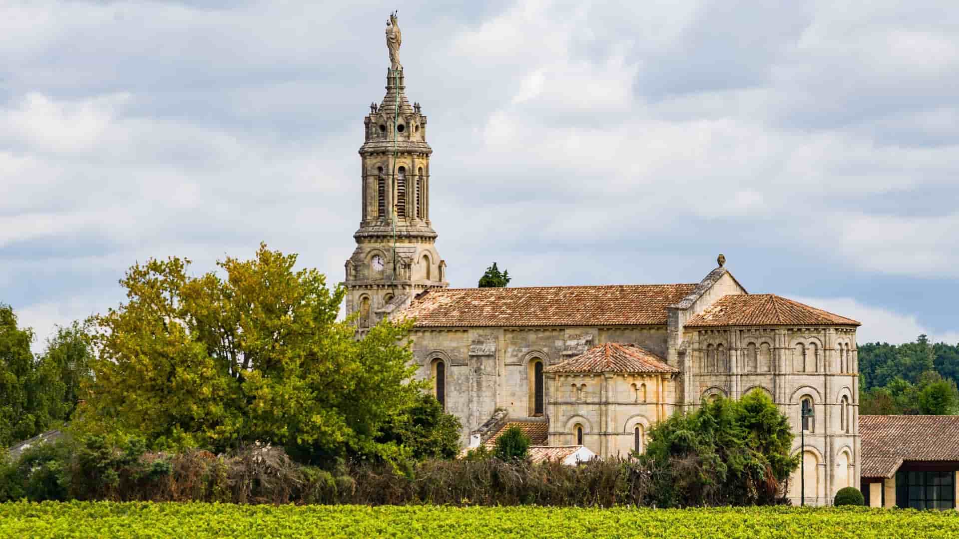 A beautiful wide-angle shot of the Église Saint-Giron church in the French town of Bourg-sur-Gironde, surrounded by a lush green vineyard under a cloudy sky.