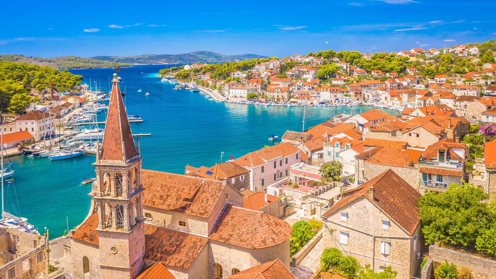 A scenic, elevated view of a coastal town on the island of Brač, Croatia. In the foreground, a historic church with a tall bell tower and a terracotta roof overlooks a bright turquoise bay. The town's buildings, with their white stone walls and red roofs, line the waterfront. The bay is filled with boats, and lush green hills covered in Mediterranean trees surround the town under a clear blue sky.