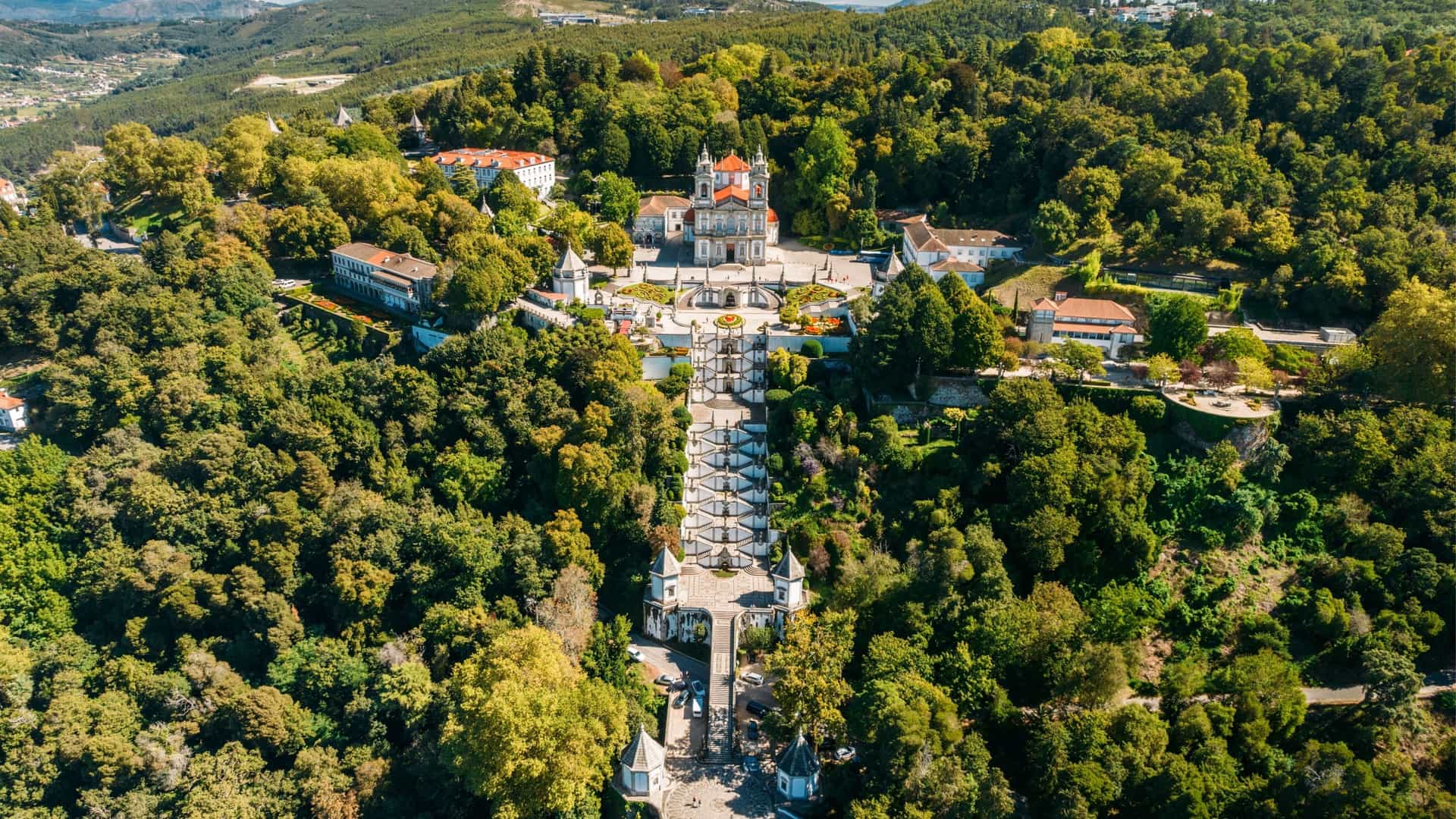 An aerial view of the Sanctuary of Bom Jesus do Monte in Braga, Portugal, featuring its famous Baroque staircase winding through a lush, forested park.
