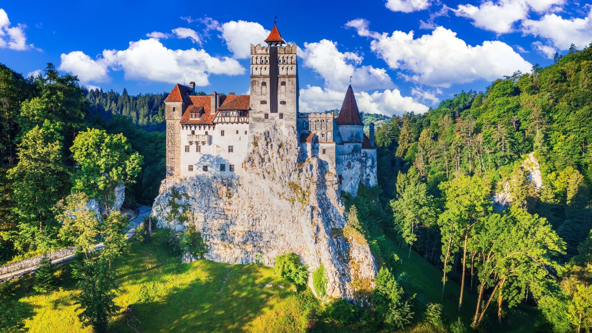 A wide-angle landscape shot of Bran Castle, a majestic landmark in Romania, often associated with the legend of Dracula, perched high on a rocky outcrop surrounded by lush forests under a vibrant blue sky with fluffy clouds.