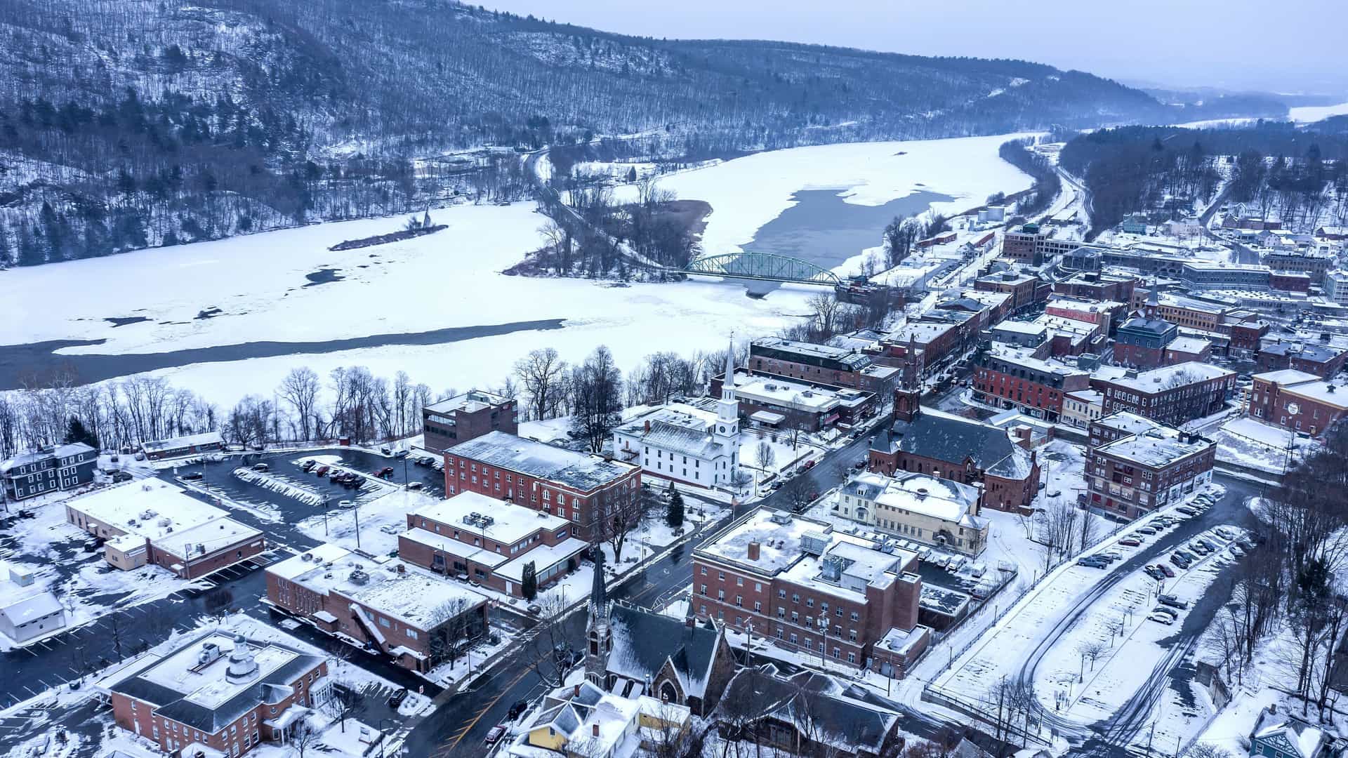 An aerial, high-angle view of the town of Brattleboro, Vermont, in winter. The rooftops and streets are covered in a fresh layer of snow, and a frozen, icy river with a bridge winds through the landscape. The town is surrounded by snow-covered, forested hills under a gray, overcast sky.