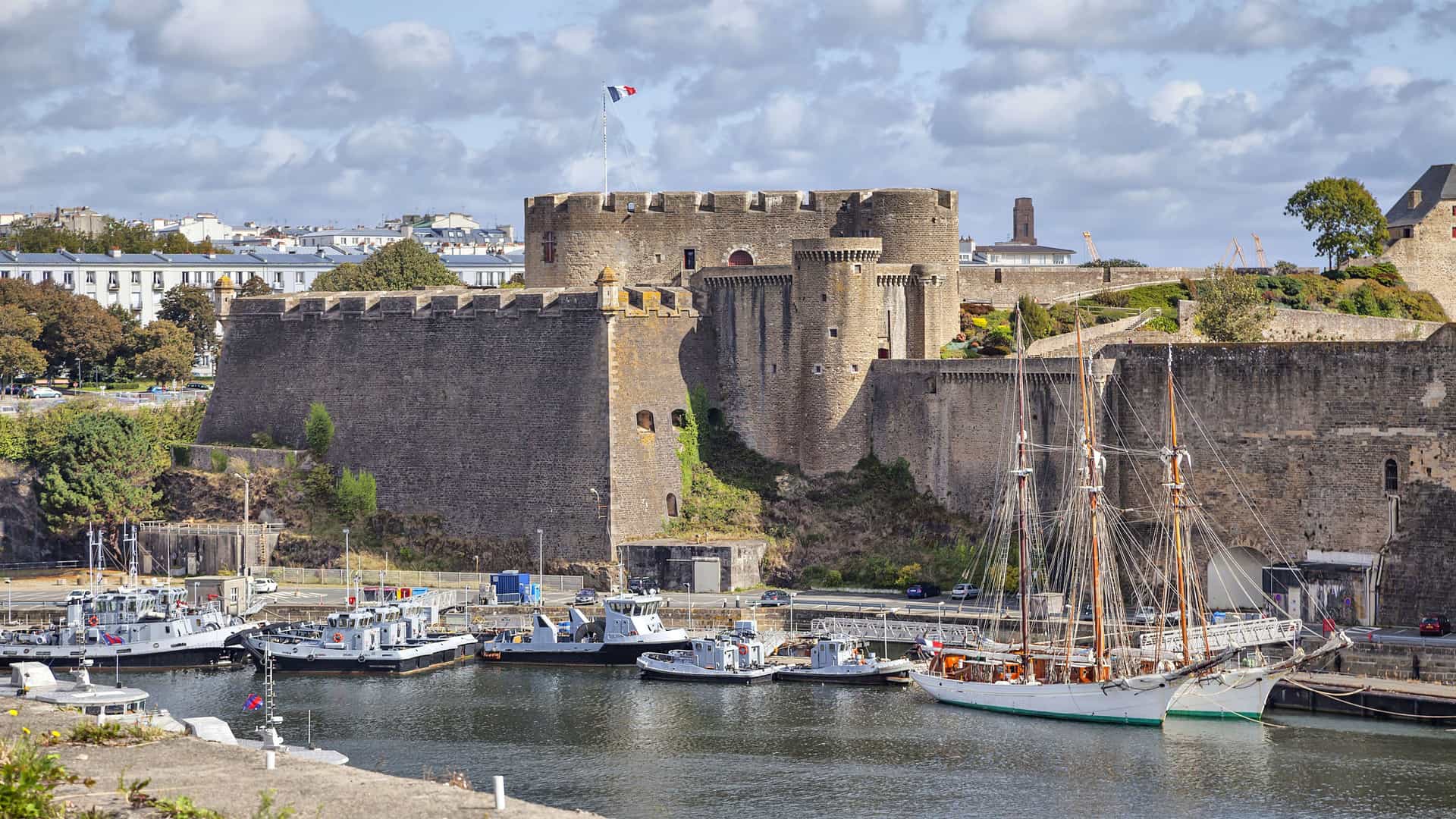 A beautiful port scene in Brest, France, with modern boats and a large sailboat docked in the foreground, and the historic medieval fortress, Château de Brest, towering over the harbor under a blue sky.
