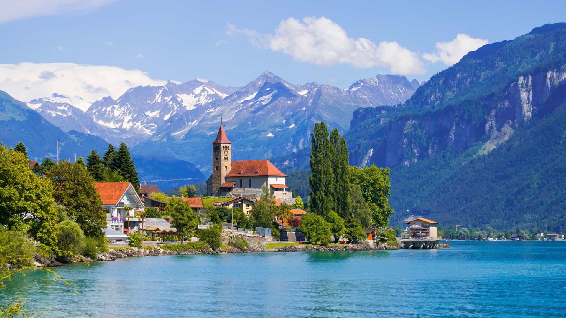 A beautiful view of Brienz, Switzerland, with the village and its church on the shore of Lake Brienz, surrounded by lush green trees and majestic, snow-capped Swiss Alps in the background.