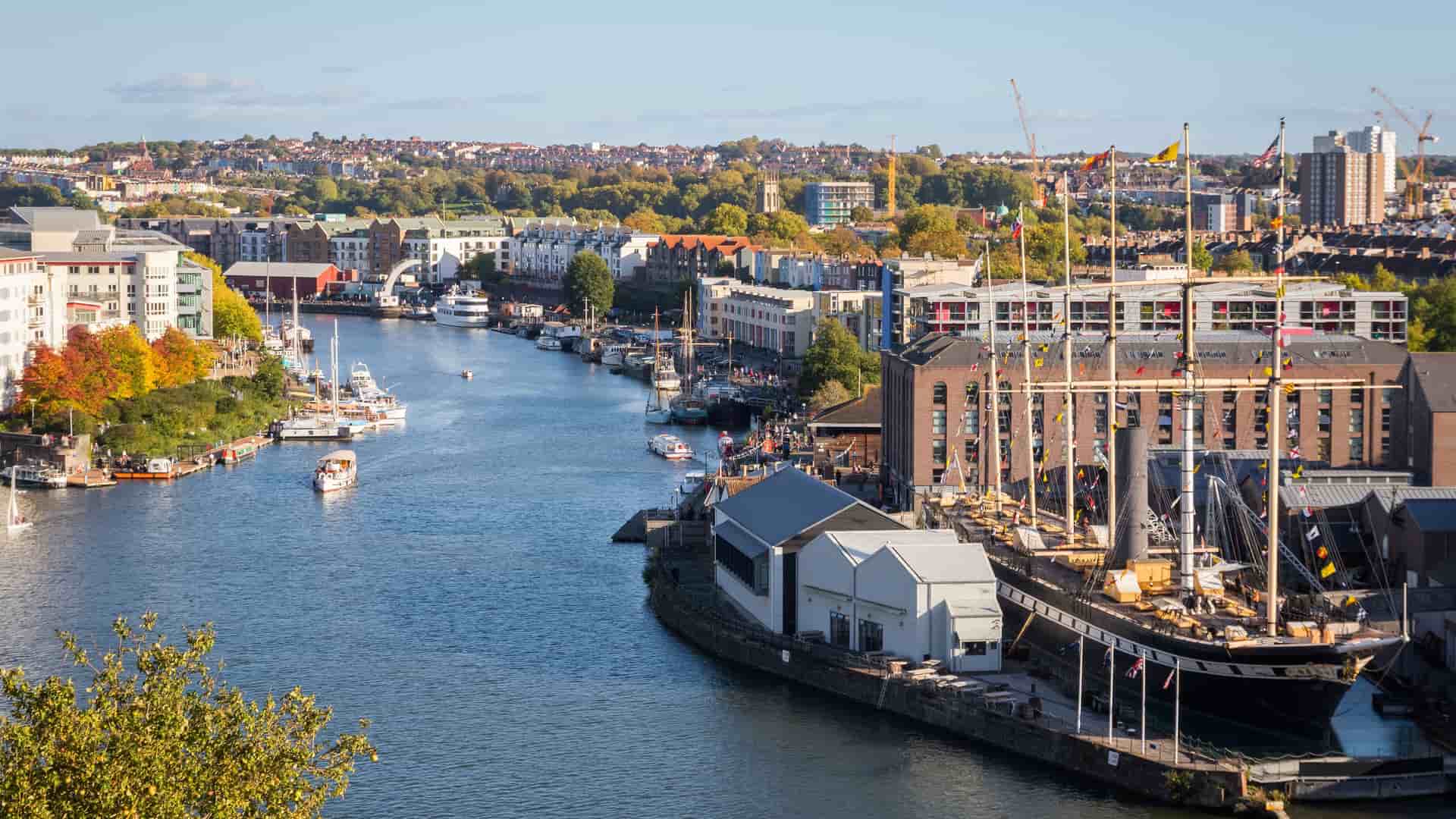 A scenic, elevated view of Bristol's Floating Harbour in England. In the foreground on the right, the large, historic sailing ship SS Great Britain is docked next to a red-brick warehouse. The water is lined with a mix of modern and older buildings, and several boats are scattered throughout the harbor. The city's landscape, with green hills and more buildings, stretches into the background under a blue sky with scattered clouds.