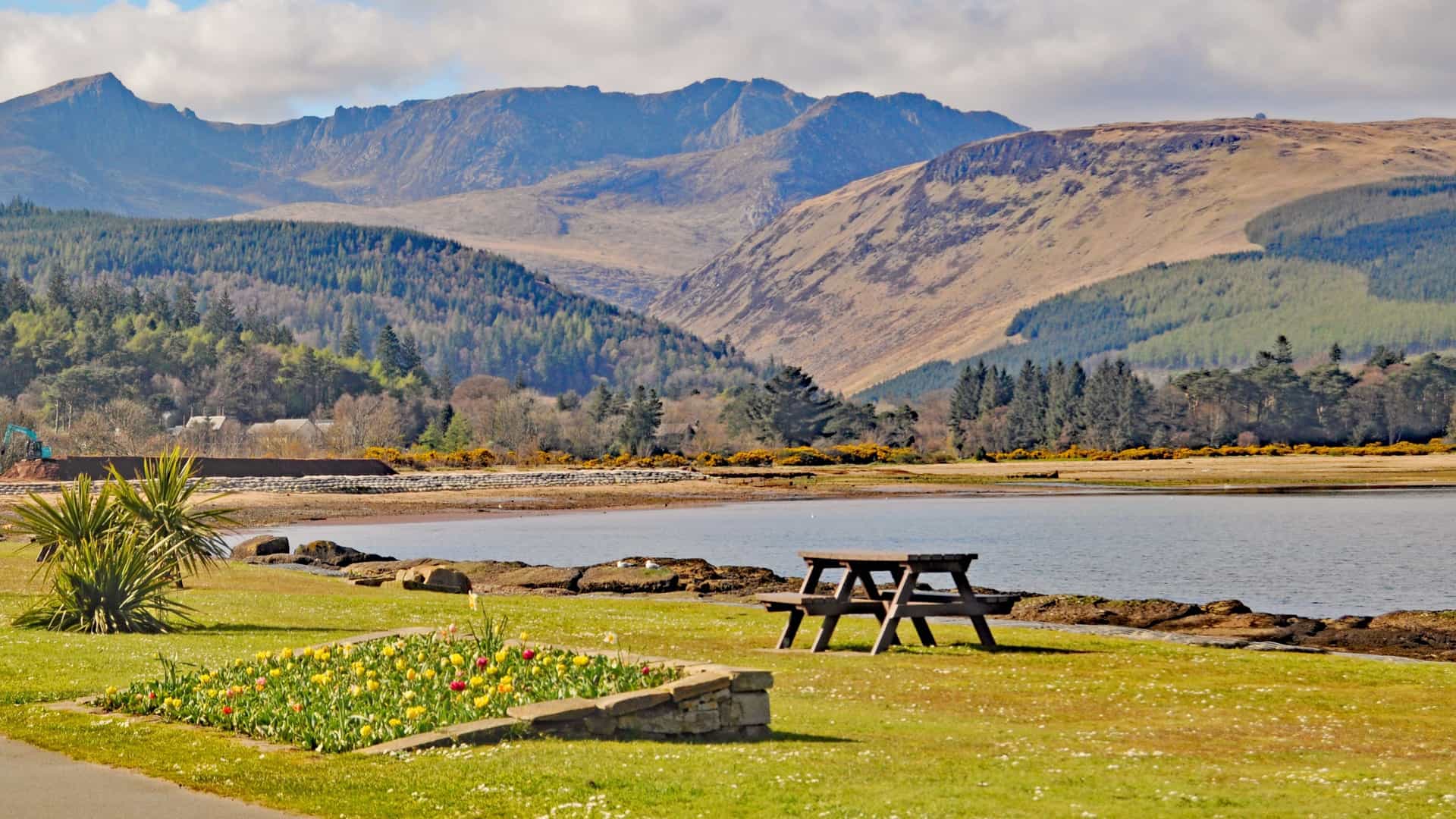 A scenic view of the Brodick Bay waterfront in Scotland, with a picnic table and a small flower bed in the foreground. The midground features the calm water of the bay, while the background is dominated by the dramatic, rugged peaks of the Goat Fell mountain range, with forested hillsides leading down to the water's edge under a partly cloudy sky.