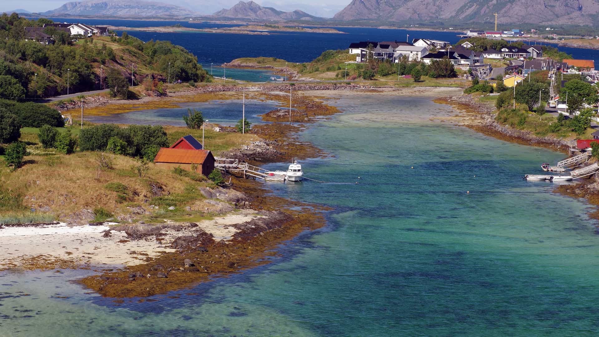 A stunning coastal landscape of Brønnøysund, Norway, with a small boathouse on the shore of a clear bay, surrounded by a charming village and rugged mountains in the distance.
