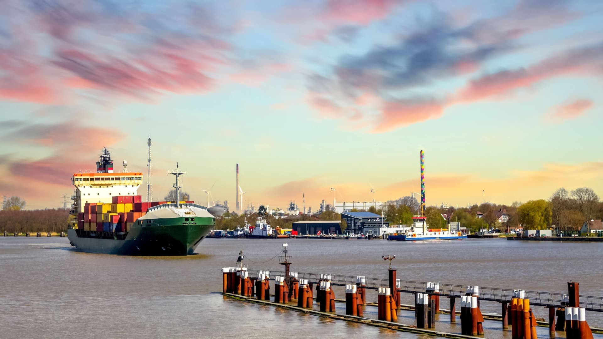A large, green container ship moves along the water near a wooden dock in the harbor of Brunsbüttel, Germany, under a colorful sunset sky.
