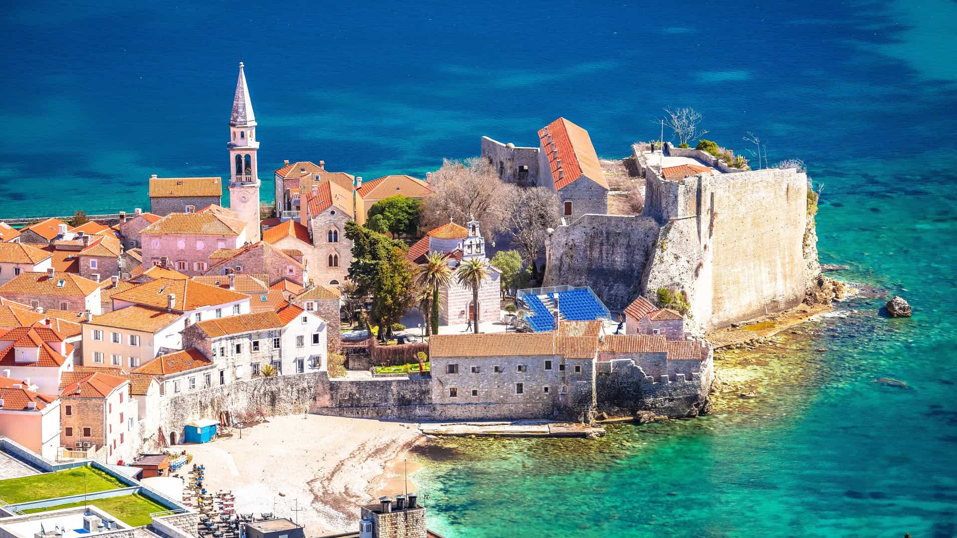 An aerial view of the historic walled town of Budva, Montenegro, situated on a small peninsula in the turquoise Adriatic Sea. The town is densely packed with buildings featuring characteristic red terracotta roofs and stone facades. A prominent church steeple rises on the left, while a large stone citadel with defensive walls stands on the right, extending into the clear blue water.