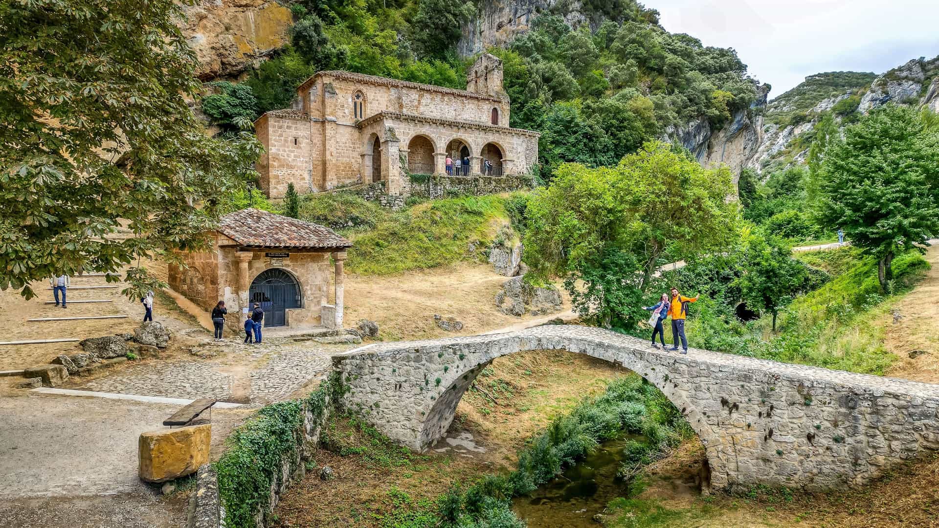 A view of the historic Hermitage of San Bernabé near Burgos, Spain, nestled into a green, rocky hillside. In the foreground, a small, ancient stone arch bridge crosses a stream. People are visible on the bridge and on the path leading to the hermitage, which is surrounded by lush trees and a smaller stone building.