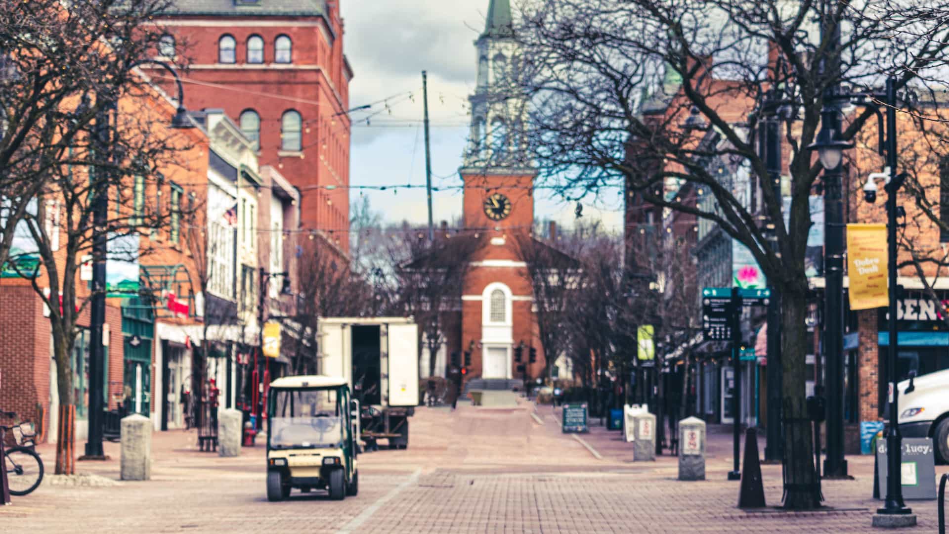 A street-level view of the Church Street Marketplace, a pedestrian-only street in Burlington, Vermont, on a cloudy day. The brick-paved street is lined with historic brick buildings and bare trees, leading to the prominent red-brick church with a clock tower and tall steeple in the background.