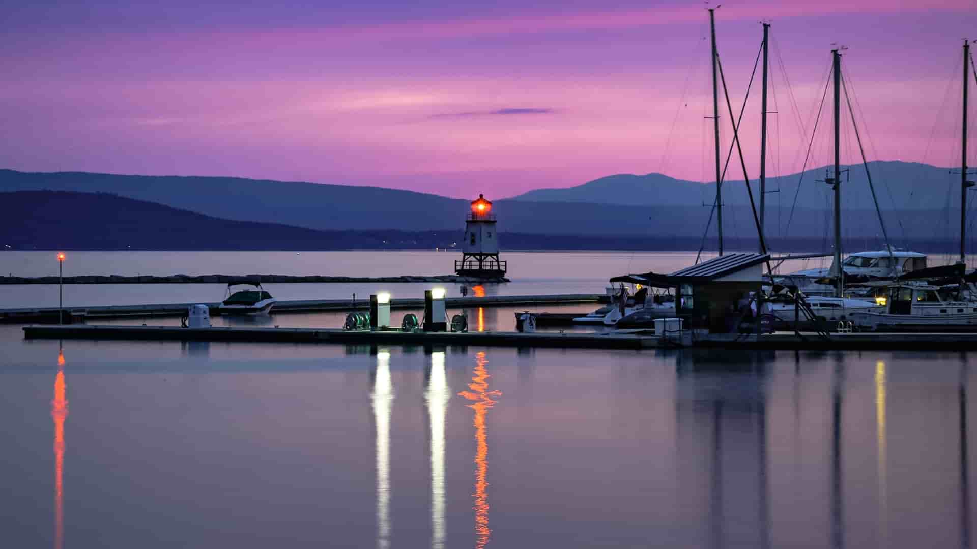 A tranquil dusk view of the Burlington harbor on Lake Champlain, with a lighthouse and sailboats at the dock.