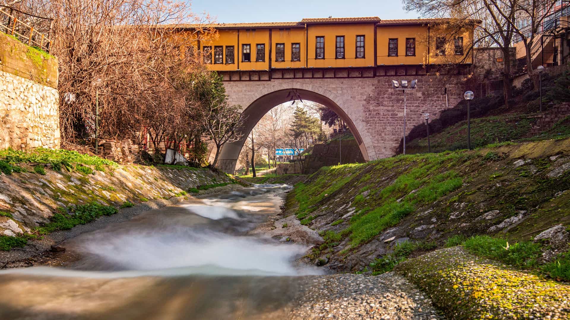 A scenic view of the historic Irgandı Bridge in Bursa, Turkey. The bridge, which has a stone arch and a long yellow building with windows on top, spans a river with flowing, blurred water. The river banks are covered in green grass and moss, with bare trees and a clear blue sky in the background.