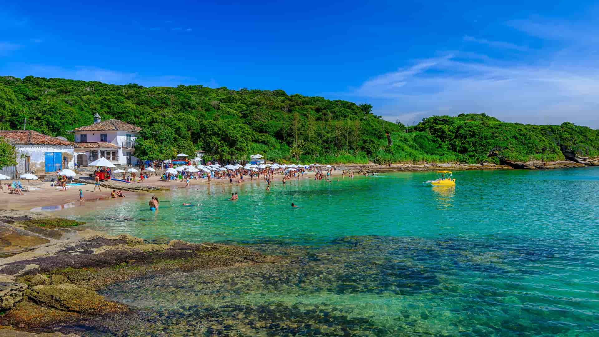 A vibrant beach scene in Búzios, Brazil, with people swimming and relaxing under white umbrellas on the sand, next to lush green hills and a small white building.