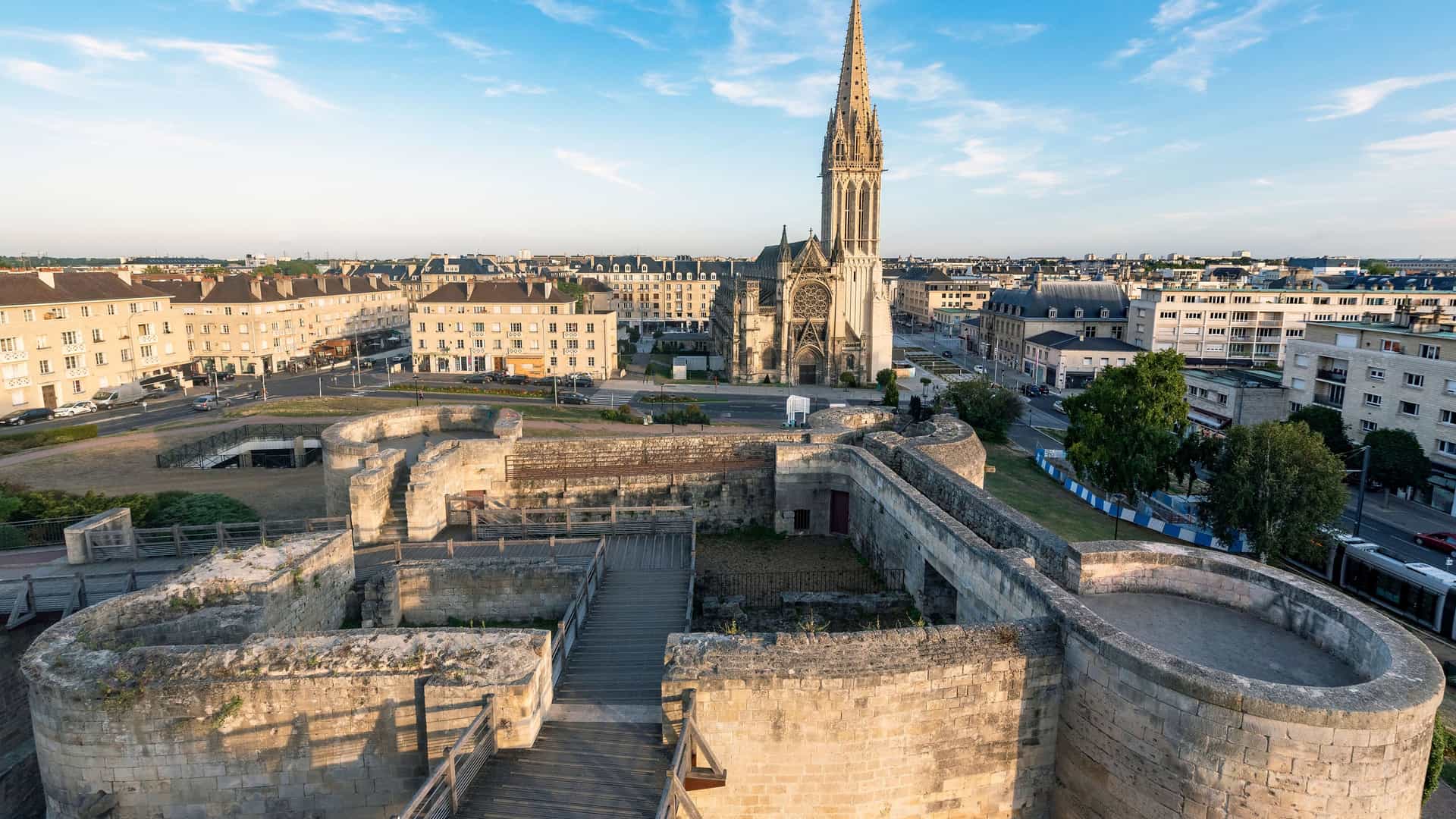 An elevated view of the historic city of Caen, France, at sunset. In the foreground are the extensive stone ruins of a medieval castle, with a wooden walkway for visitors. In the background, the ornate, towering spire of the Église Saint-Pierre rises above the surrounding urban landscape of modern and historic buildings under a clear blue sky.