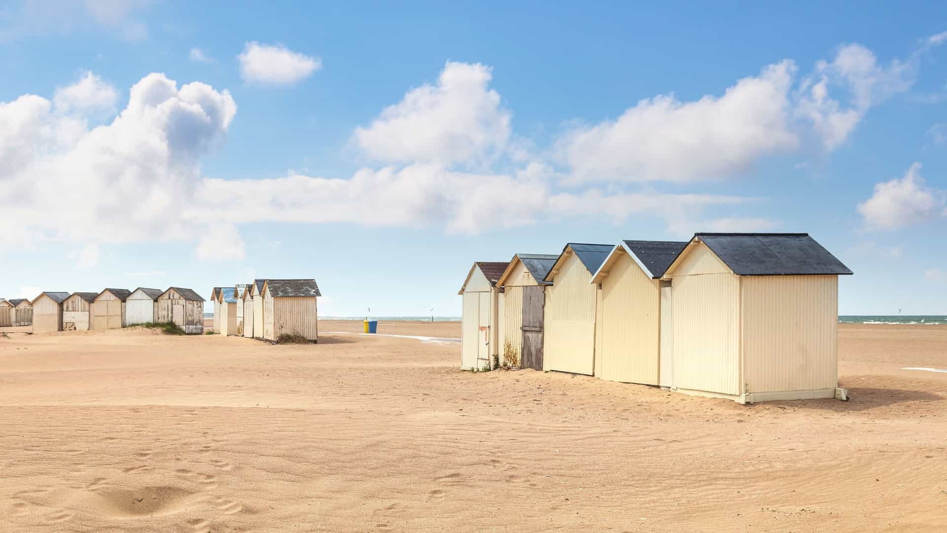 A row of traditional wooden beach huts on the sandy shores of Ouistreham, France, with a blue sky and white clouds overhead.
