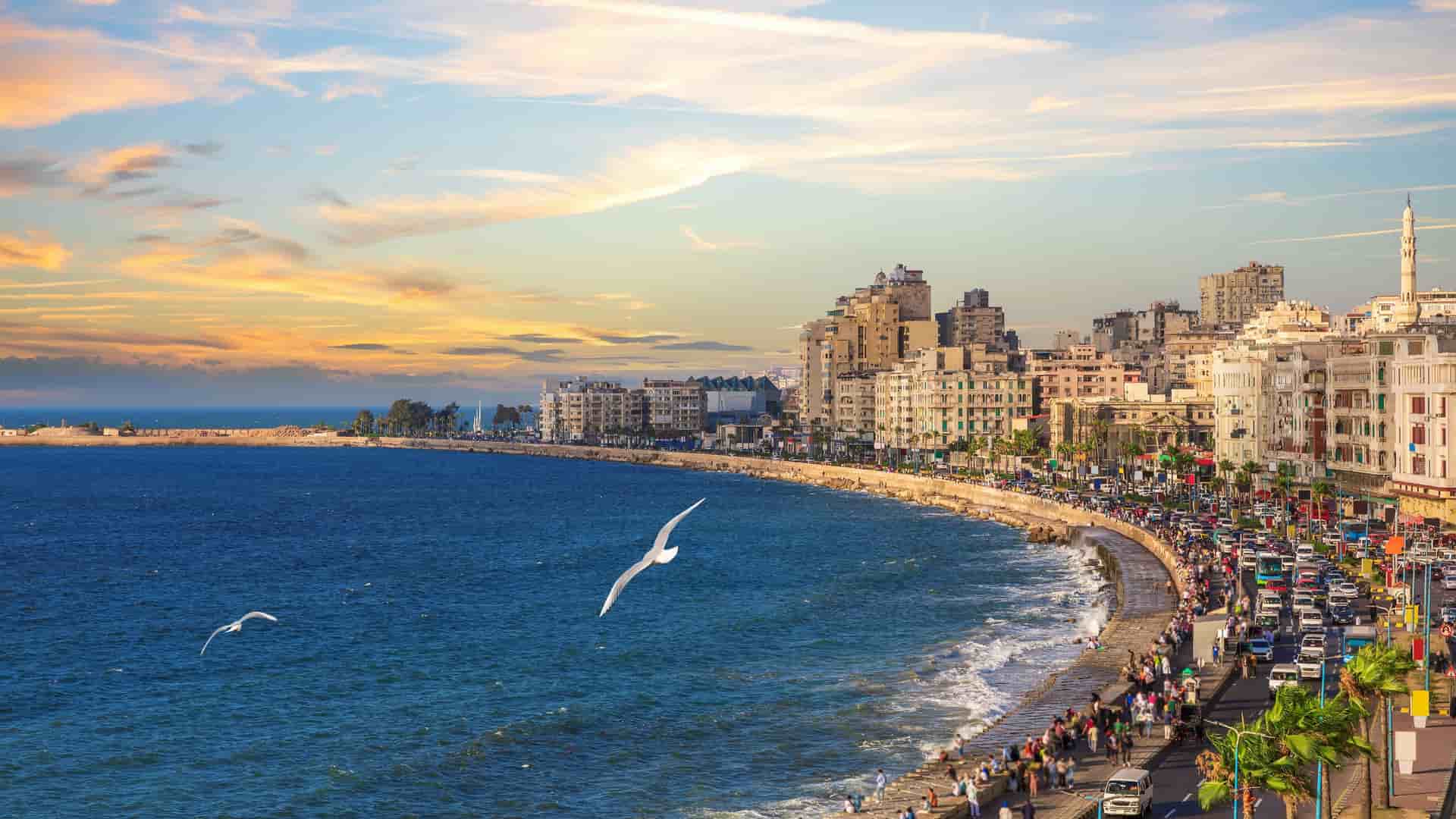 A vibrant wide-angle view of the bustling city of Alexandria, Egypt, with its urban coastline curving along the Mediterranean Sea at sunset.