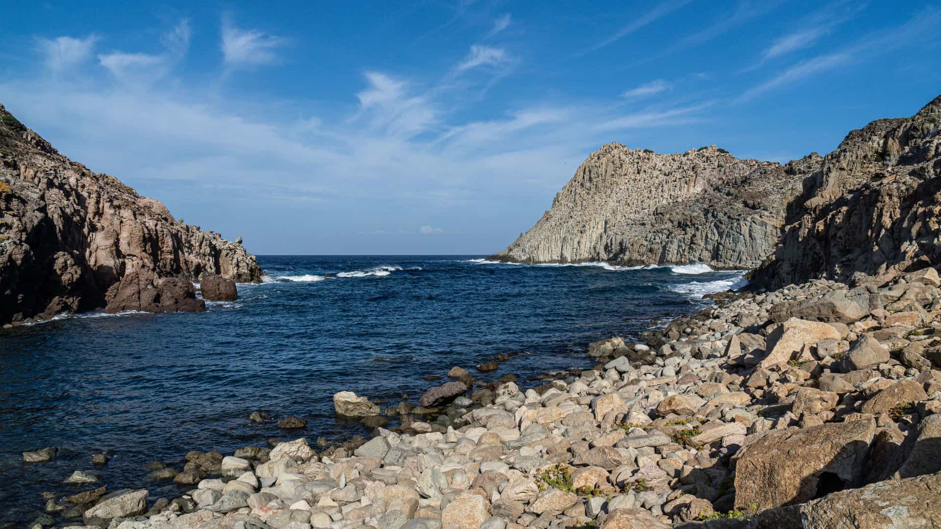 A scenic view of Cala di Fico on the island of Capraia, Italy, with a rocky coastline, turquoise water, and a dramatic cliff formation under a blue sky.