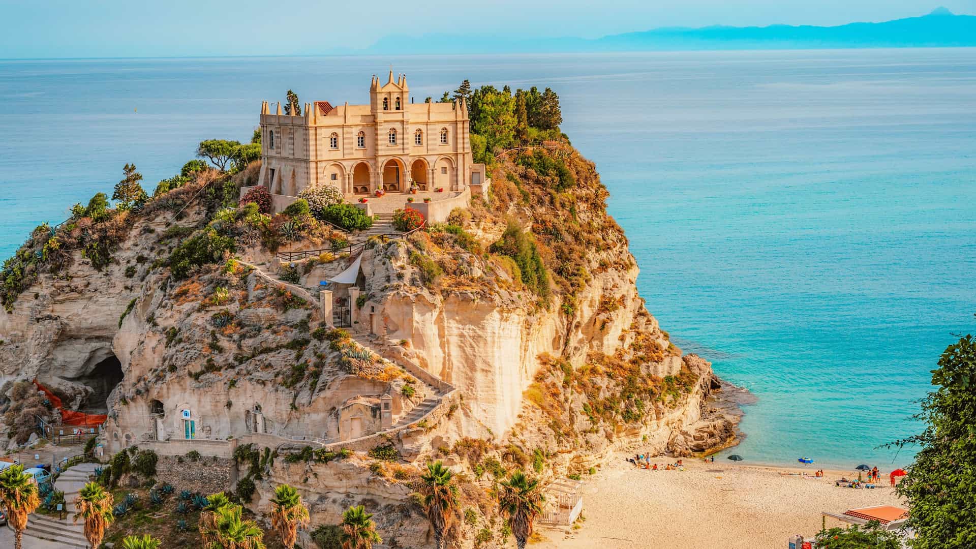 A scenic view of the Sanctuary of Santa Maria dell'Isola, a medieval monastery, perched on a rocky cliff overlooking a sandy beach and the turquoise waters of the Tyrrhenian Sea in Tropea, Calabria, Italy. Stone stairs are carved into the cliffside, and people are visible on the beach below.