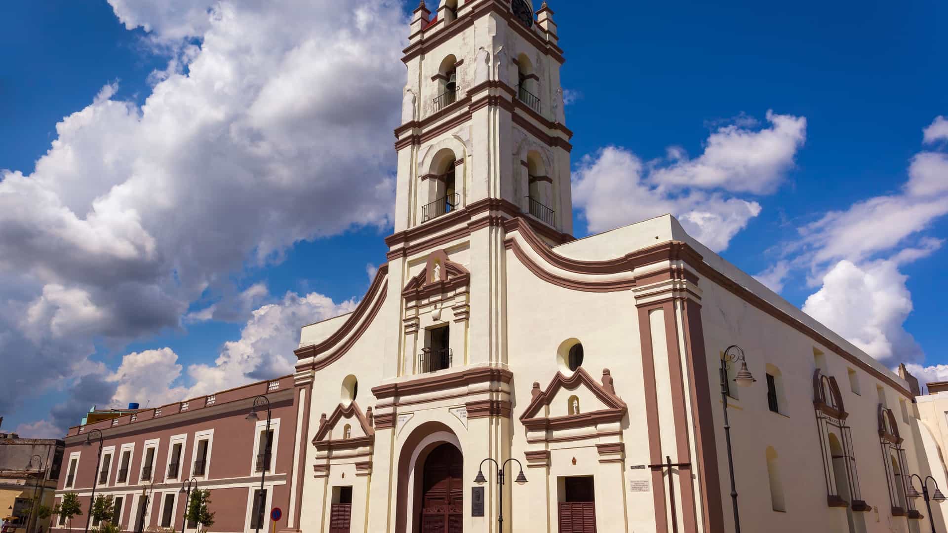 The historic Iglesia Parroquial de Nuestra Señora de la Soledad in Camagüey, Cuba, a colonial-style church with a prominent bell tower and distinctive reddish-brown trim, stands under a vibrant blue sky with large white clouds.