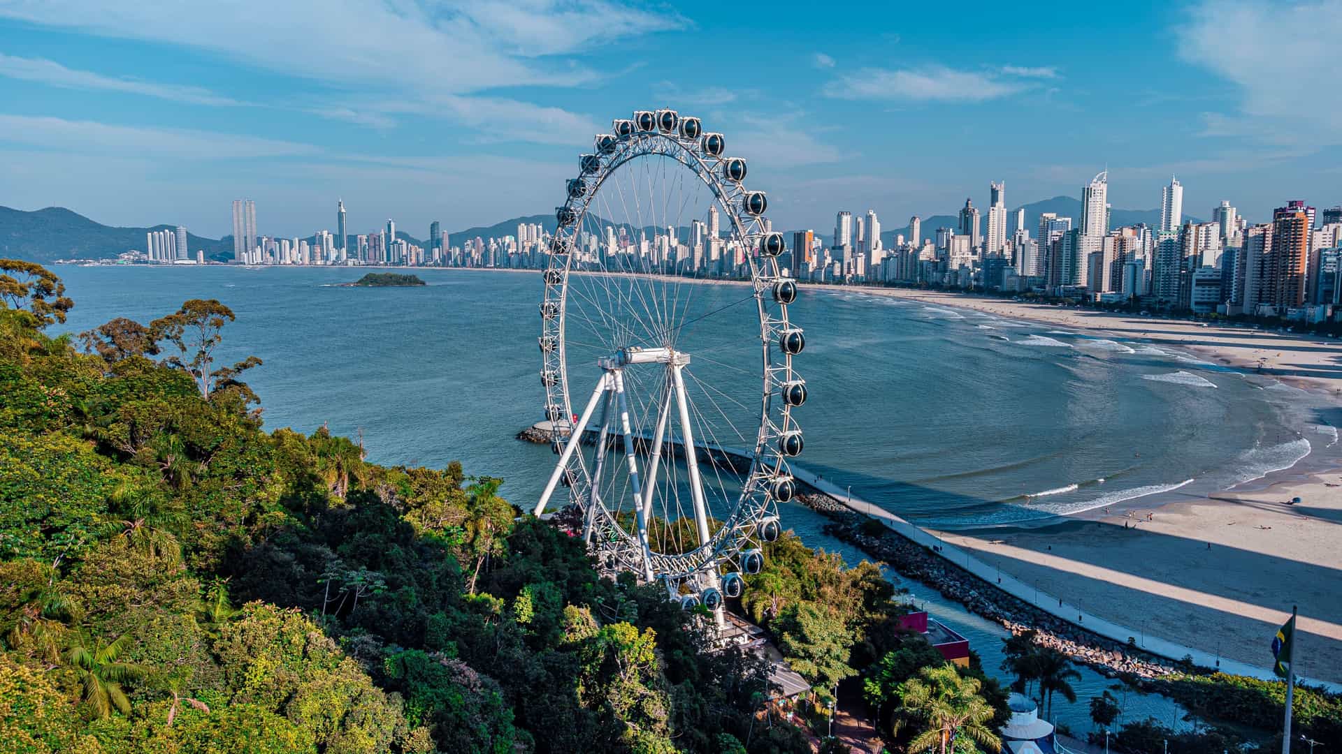 An aerial view of the Balneário Camboriú Ferris wheel, a landmark attraction on the Brazilian coastline, with the city's impressive skyline and sandy beach in the background.