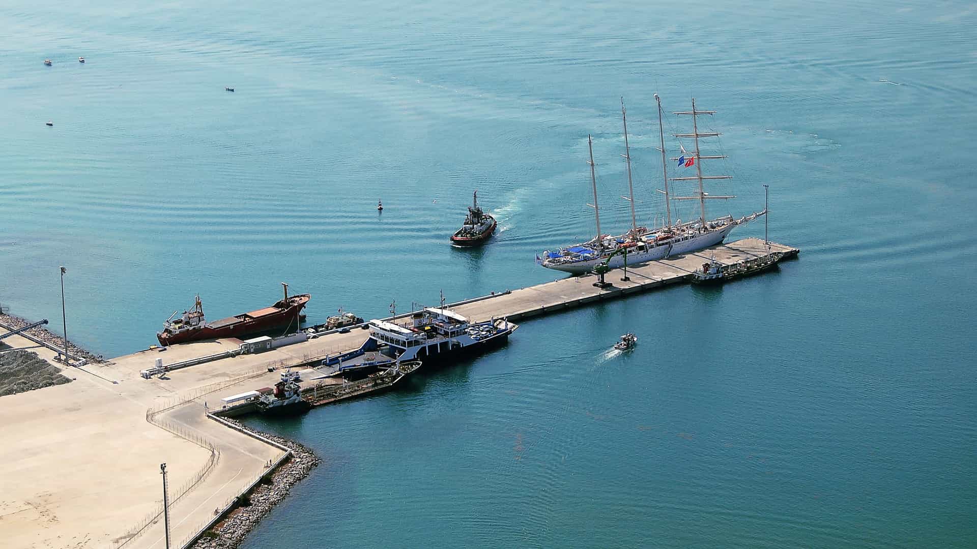A busy harbor scene in Canakkale Kepez with a long pier lined by multiple vessels, including a large red cargo ship, a tall‑masted sailing ship, and smaller boats, while a tugboat moves through calm water creating ripples against the backdrop of port infrastructure.