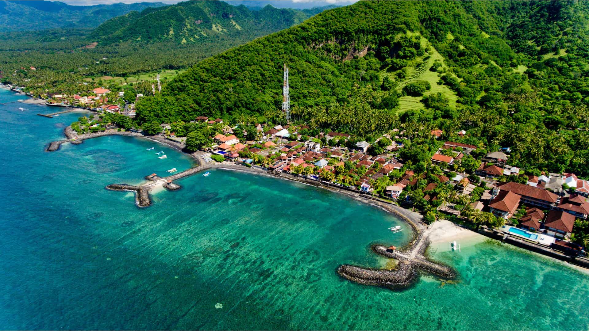An aerial view of the coastal town of Candidasa, Indonesia, nestled along a series of coves with vibrant turquoise water. Stone breakwaters extend into the clear water, while the town's buildings, with their red-tiled roofs, are set among lush green tropical foliage at the base of a steep, verdant hillside.