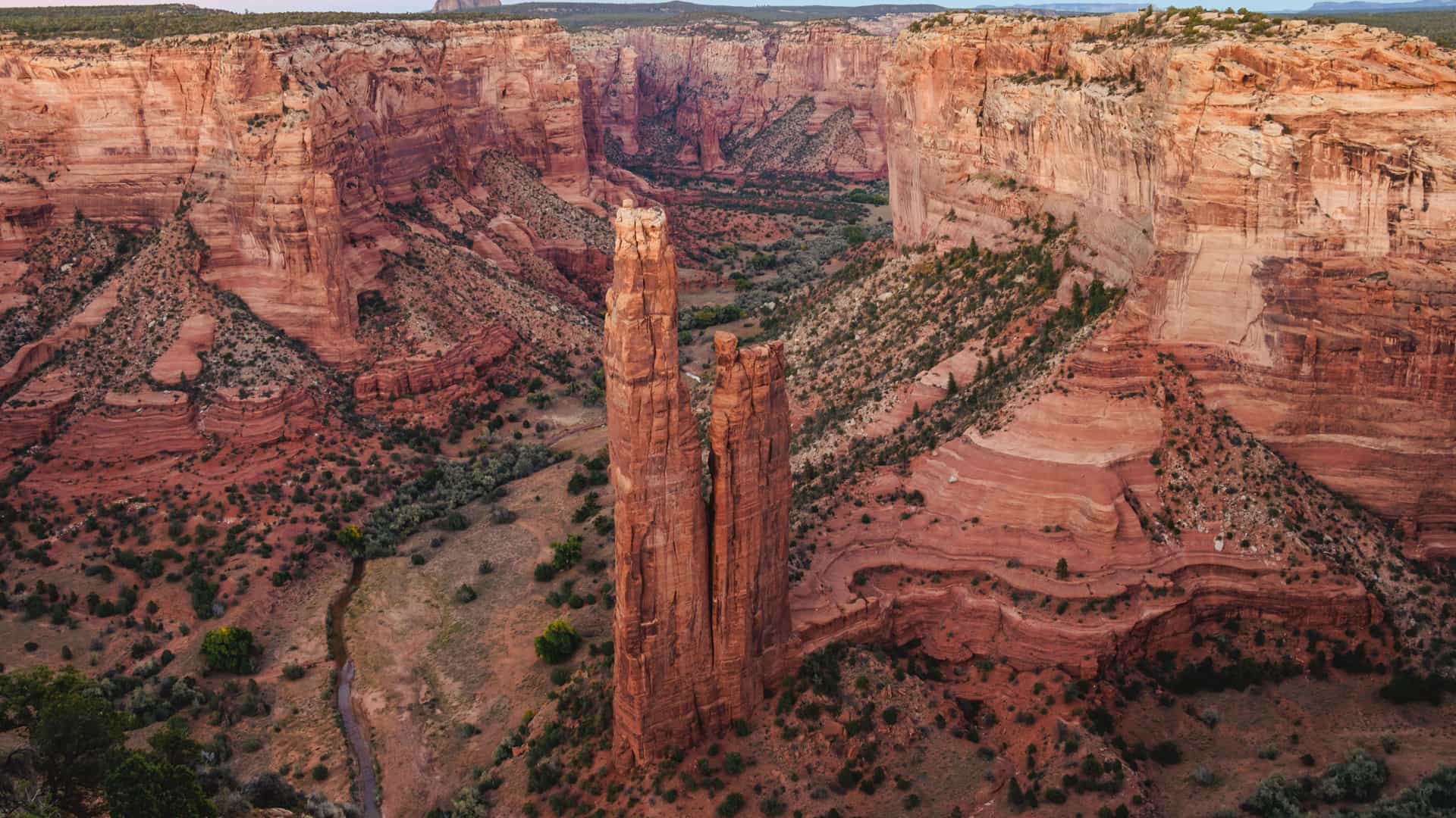 A panoramic, high-angle view of the iconic Spider Rock spire in Canyon de Chelly National Monument, Arizona. The towering red sandstone formation rises from the canyon floor, which is bisected by a winding stream and covered in green trees. The canyon walls show striated layers of red and orange rock under a soft, warm-colored sky.
