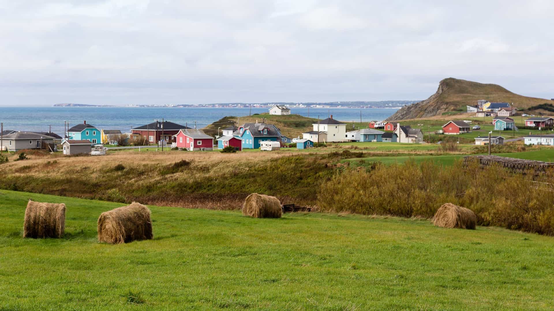 A panoramic landscape of the rolling green hills and hay bales of Cap-aux-Meules, Quebec, with a cluster of colorful houses overlooking the sea and a small town in the distance.