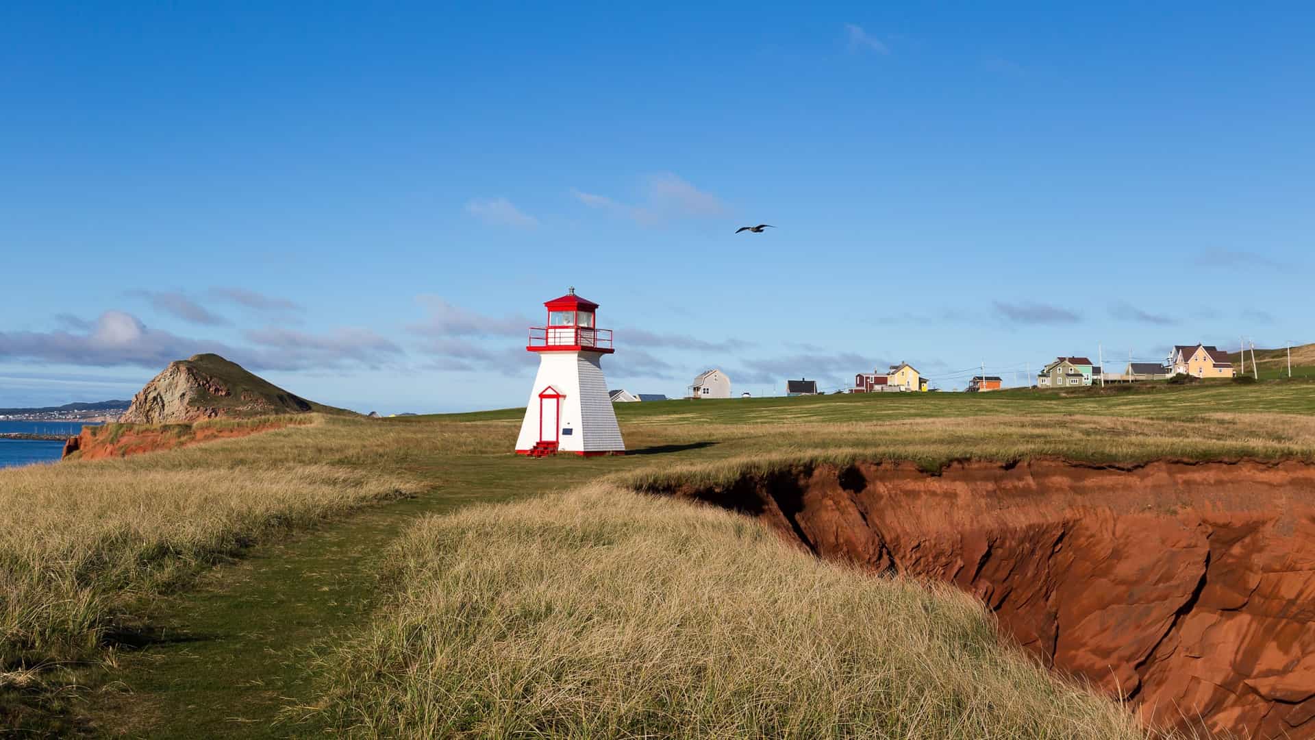 A charming white and red lighthouse, perched on a dramatic red cliff overlooking the ocean, with lush green grass and a few houses under a bright blue sky on Cap-aux-Meules, Quebec.