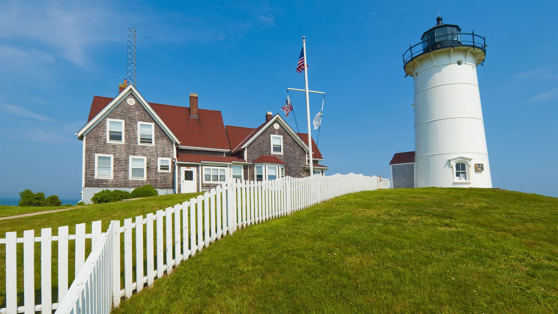 A beautiful daytime view of the iconic Nobska Lighthouse, a white lighthouse with a black top, and the attached keeper's house on a lush green hill with a white picket fence on Cape Cod, Massachusetts.