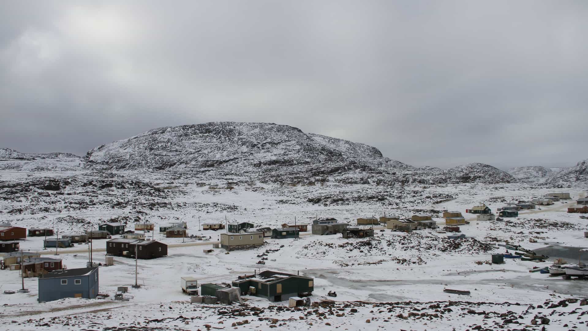 A winter landscape shot of the remote Inuit community of Cape Dorset, also known as Kinngait, with small, colorful houses nestled in the snow-covered Arctic tundra under a grey sky.