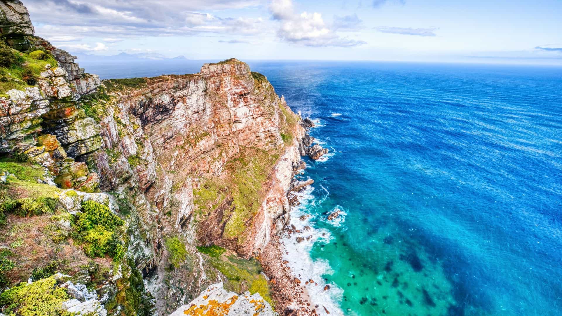 A breathtaking view from the cliffs of Cape Point, South Africa, overlooking the turquoise ocean where the Atlantic and Indian Oceans meet, with waves crashing against the rocky shore.