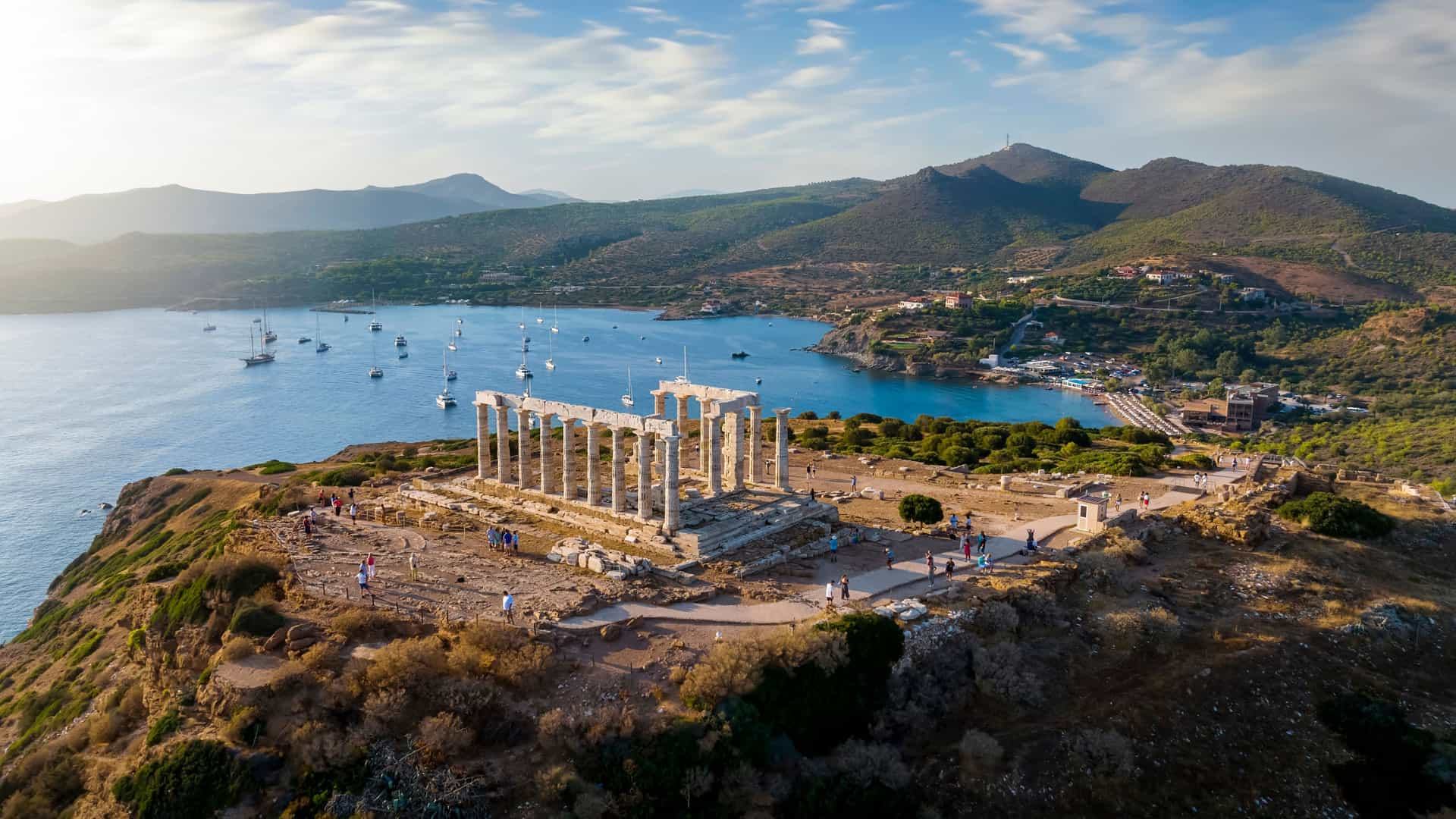 An elevated view of the ancient ruins of the Temple of Poseidon at Cape Sounion, Greece. The weathered white marble columns are perched on a rugged cliff overlooking the blue waters of the Aegean Sea, which is dotted with boats. The rocky coastline and distant hills are visible in the background under a blue sky with scattered clouds.