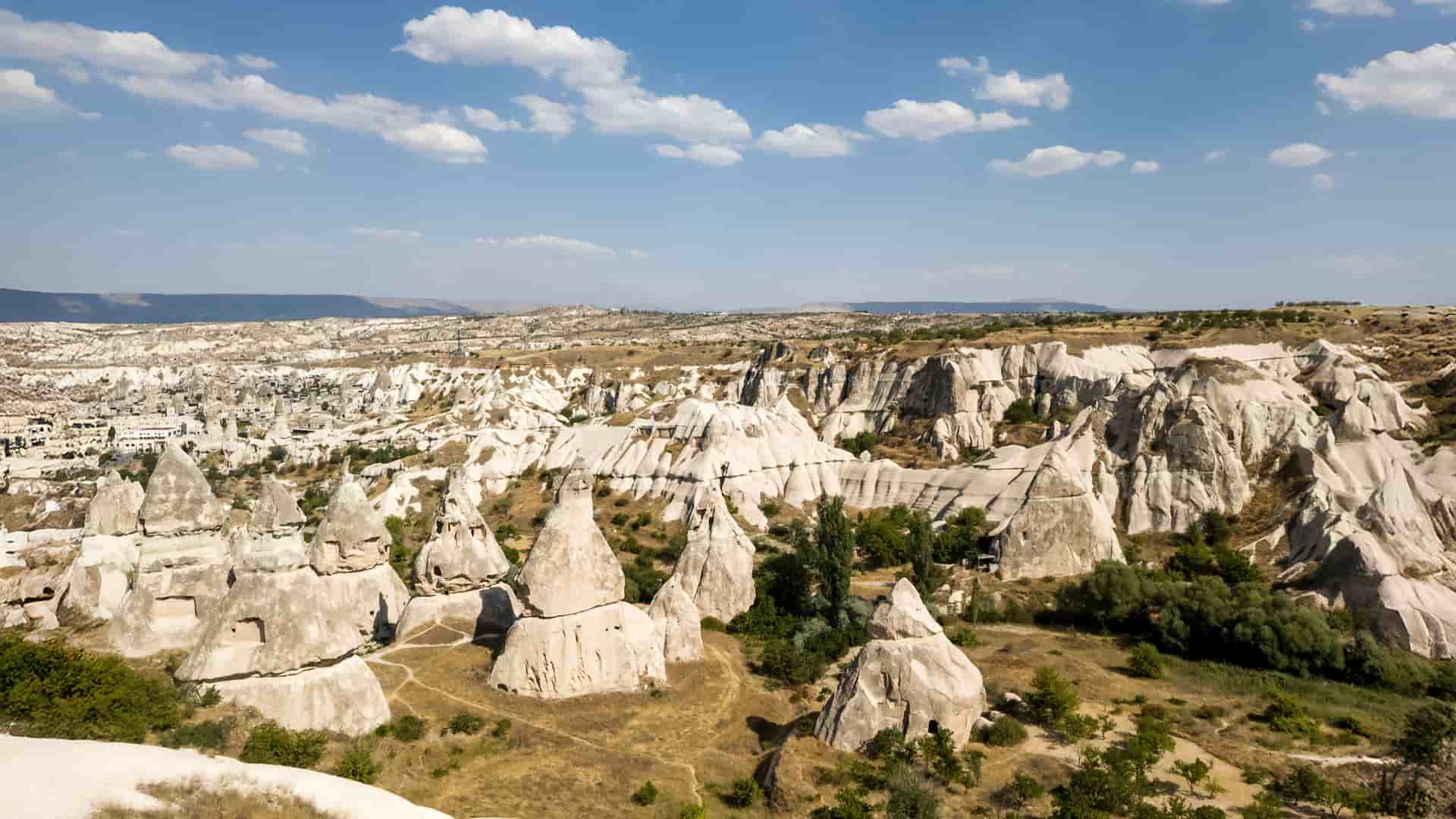 A stunning landscape of Cappadocia, Turkey, featuring unique rock formations and fairy chimneys under a bright blue sky with scattered clouds.