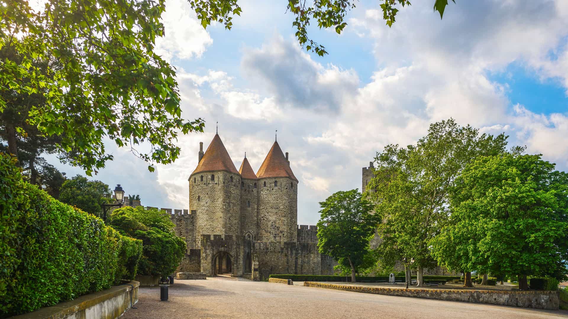 A close-up view of the main entrance to the medieval fortified city of Carcassonne, France. The stone walls and two prominent conical towers with reddish-orange roofs are framed by lush green trees and manicured hedges, all under a dynamic sky with white clouds.