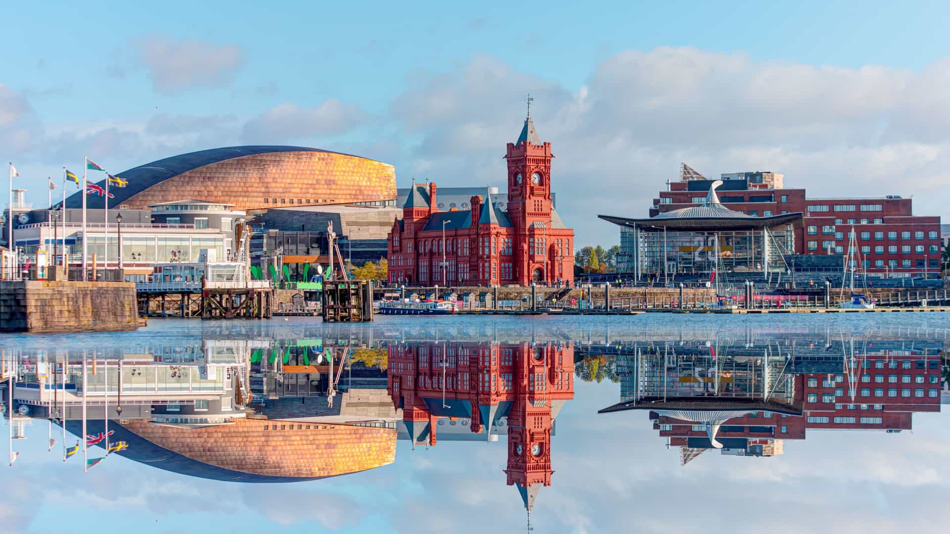 A panoramic view of Cardiff Bay with the historic Pierhead Building, the modern Wales Millennium Centre, and other landmarks reflecting beautifully on the still water.