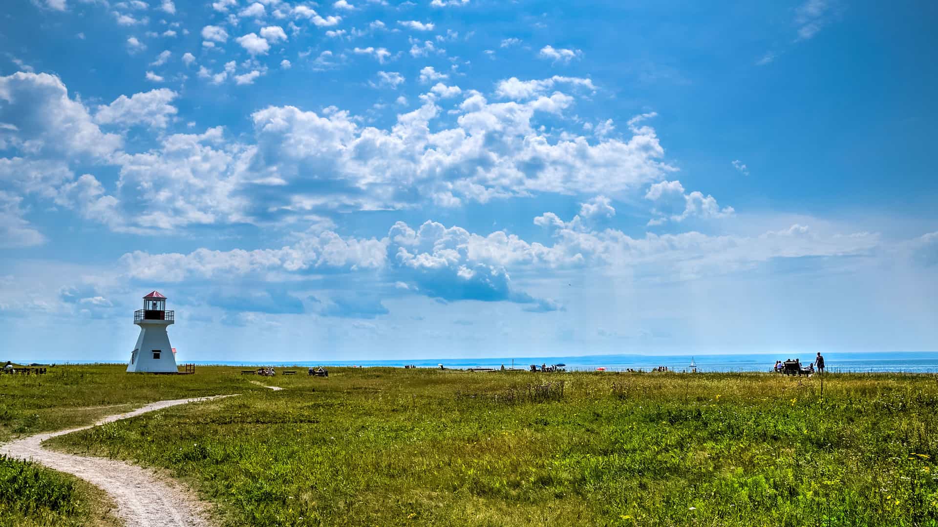 A small, white lighthouse with a red roof stands in a wide, grassy field in Carleton-sur-Mer, Quebec, Canada. A winding dirt path leads toward the lighthouse, with the calm waters of the Baie des Chaleurs visible in the distance under a bright blue sky filled with large, puffy white clouds.
