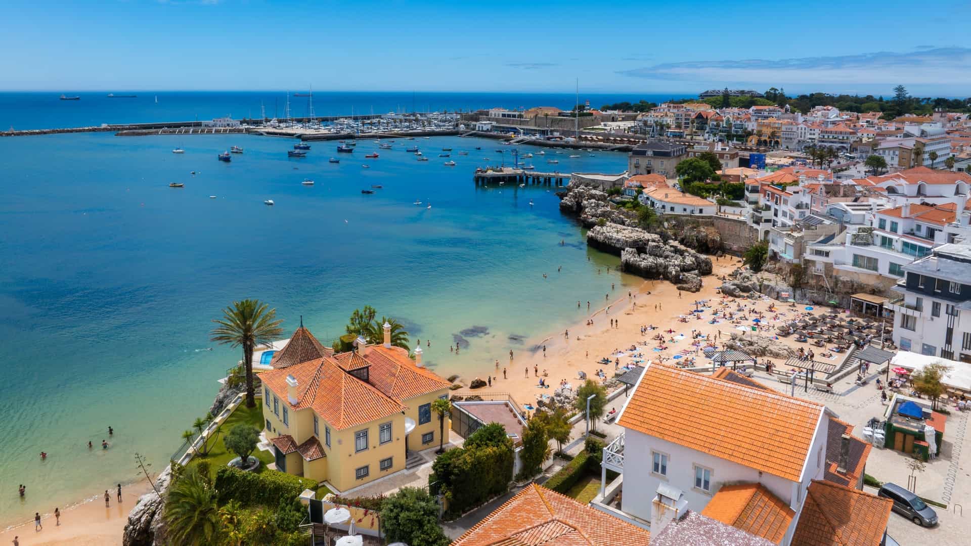 An aerial, panoramic drone view of Cascais, a coastal town on the Portuguese Riviera located about 30km west of Lisbon. The image features the crescent-shaped sandy beach and the iconic Bay, lined with the historic center's dense cluster of white and terracotta-roofed buildings. The turquoise waters of the Atlantic Ocean stretch out in the background under a bright, clear sky. The view captures the town's coastline and a section of its marina or harbor.