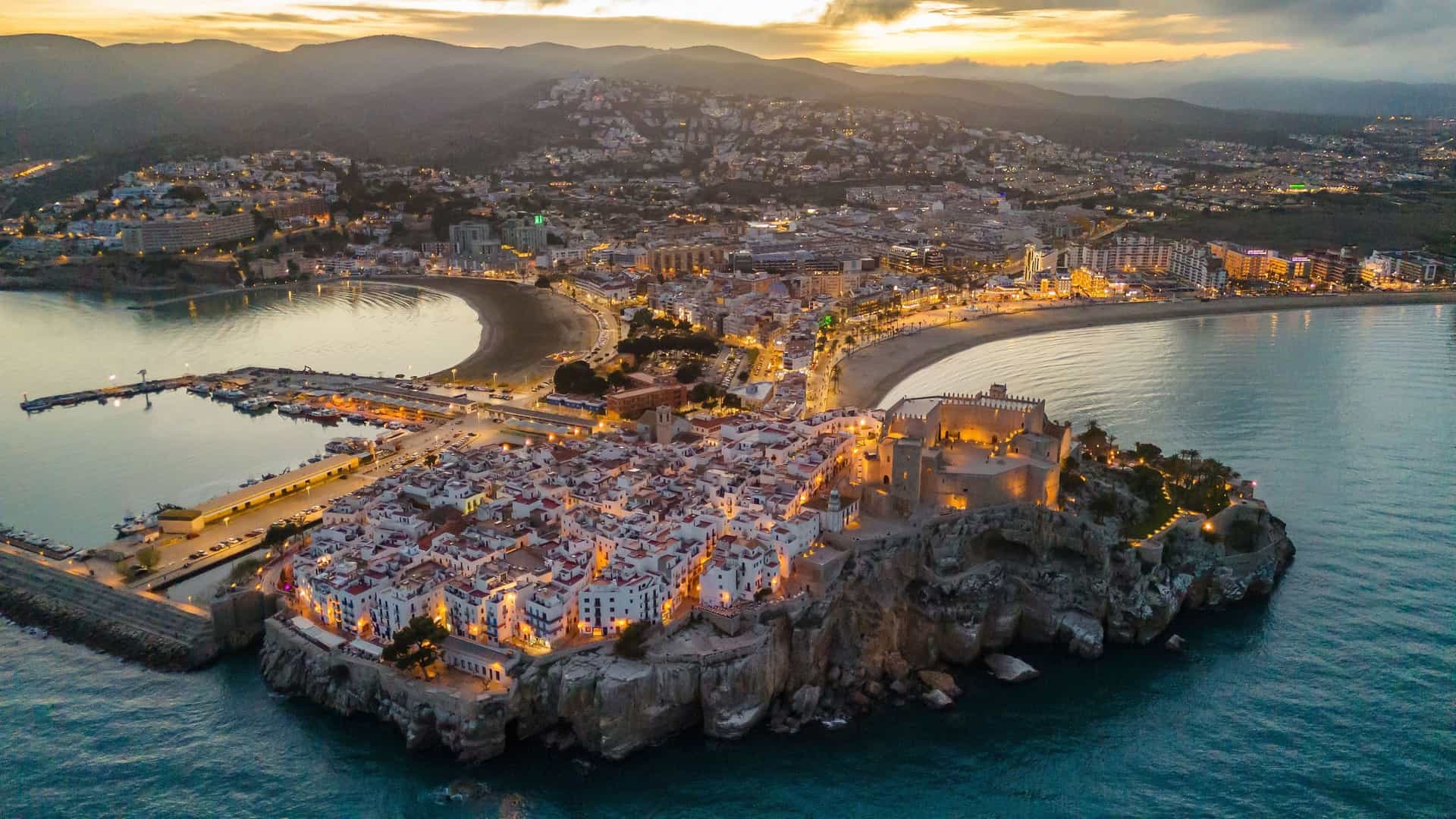 An aerial shot of Peñíscola, Castellón, Spain, at dusk, showing the illuminated Pope Luna Castle on a rocky peninsula extending into the sea.
