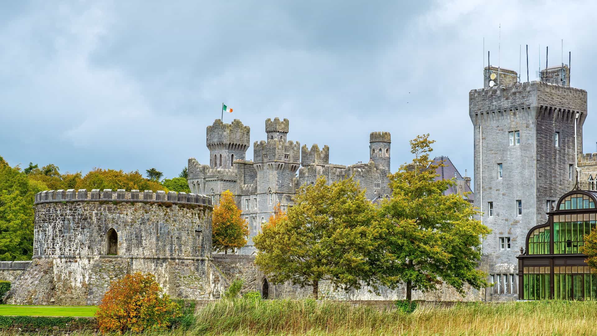 The historic Ashford Castle, a gray stone fortress with multiple towers and turrets, stands under a cloudy sky in County Mayo, Ireland. An Irish flag flies from one of the highest towers. The castle is partially obscured by lush green and autumn-colored trees, with a green lawn and modern glass structures visible in the foreground.